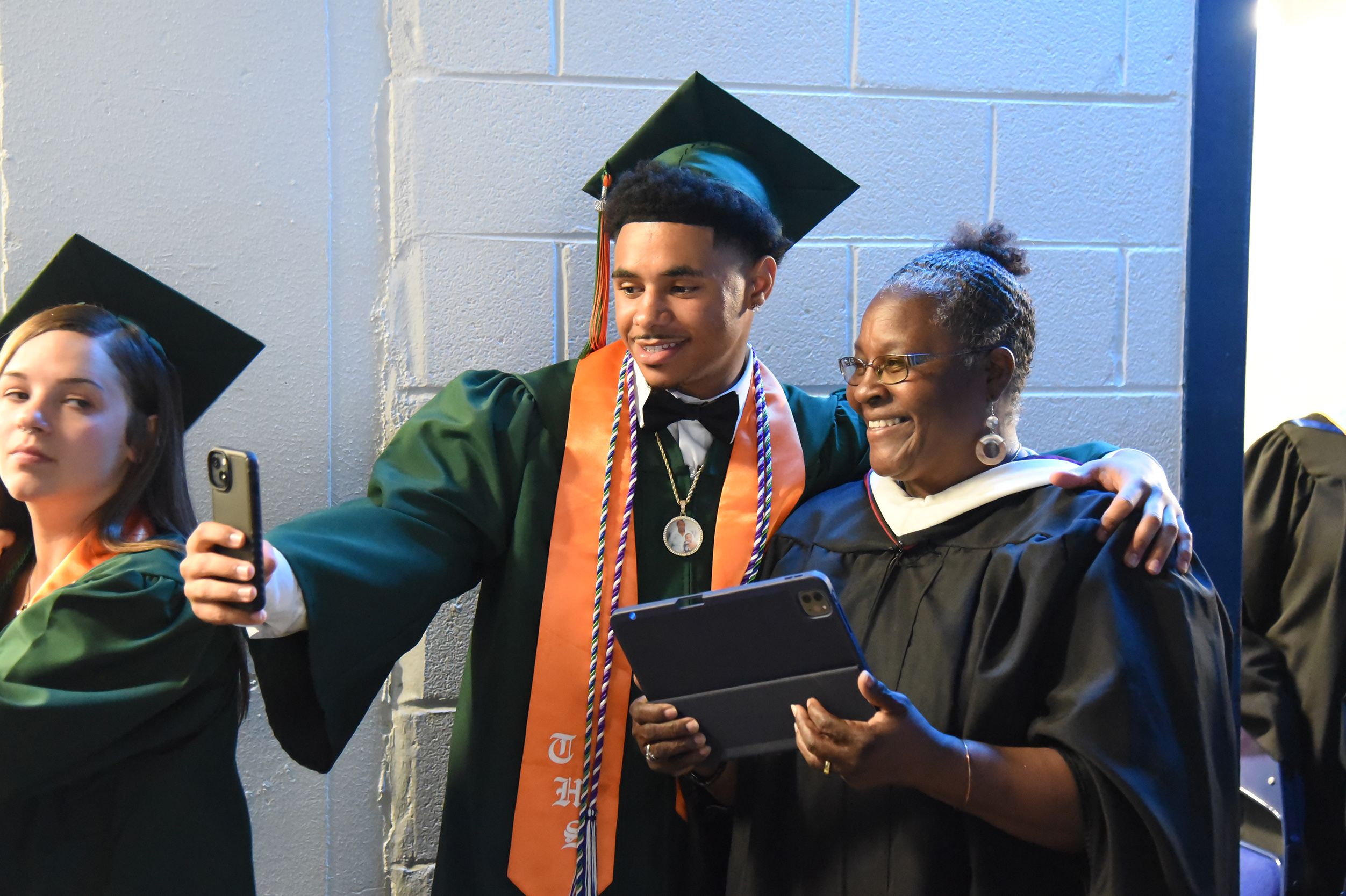 A person in a green gown and bow tie holds a tablet while a woman in black holds it.