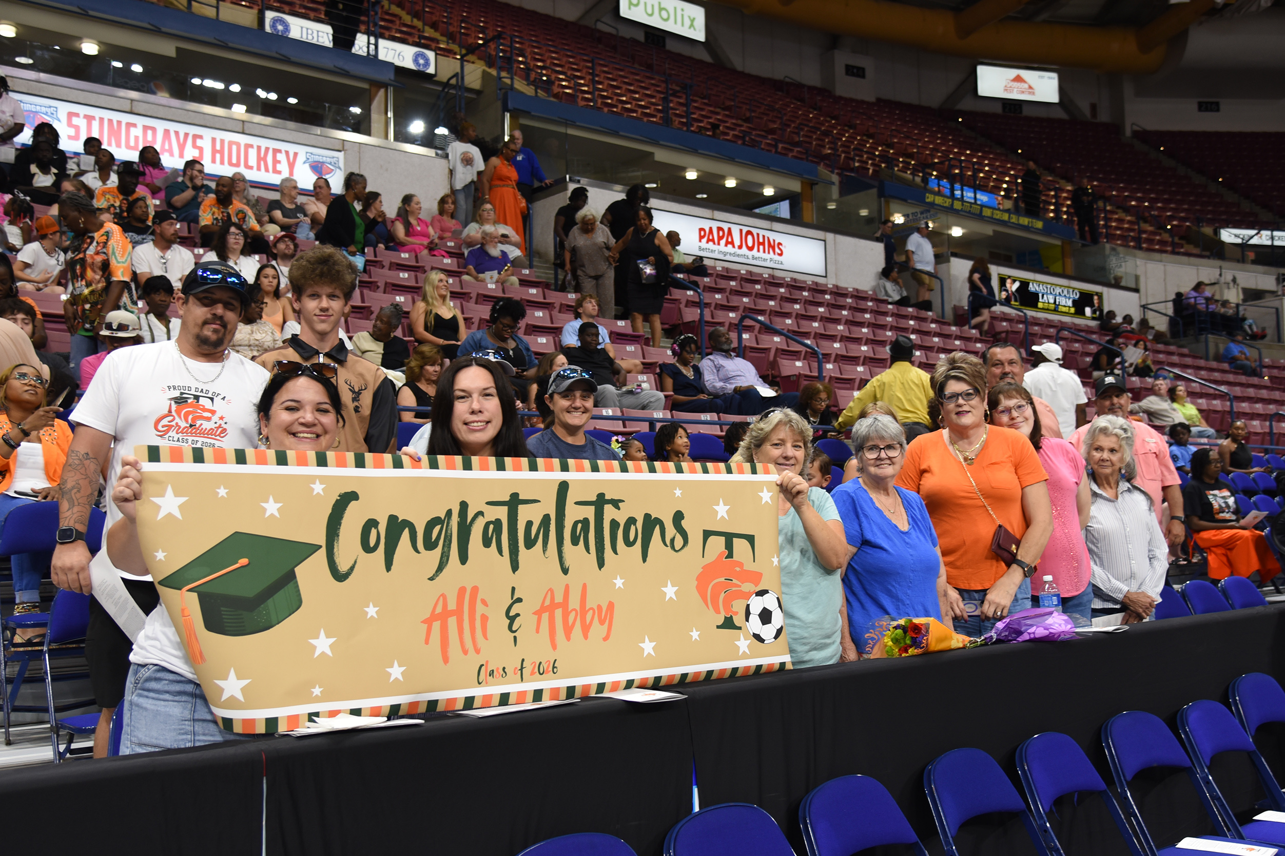 People in a stadium, some wearing caps, holding a banner with "Congratulations Ali & Abby" and a cap illustration.