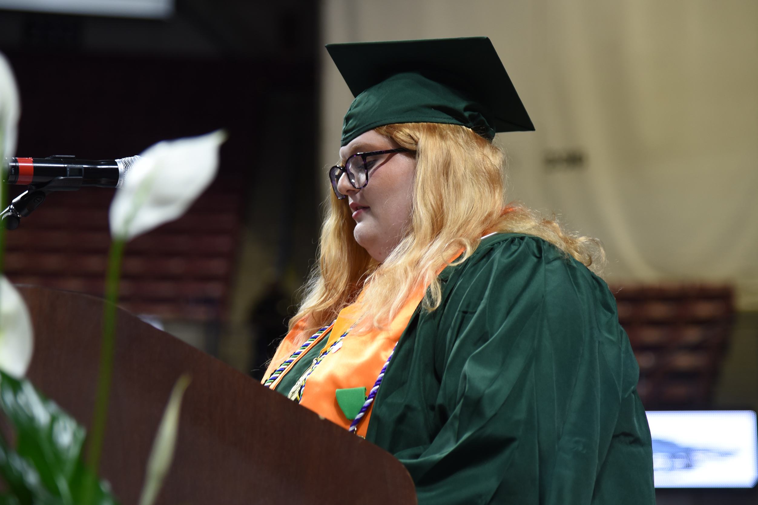 A woman wearing a green graduation gown and cap speaks into a microphone at a graduation ceremony.