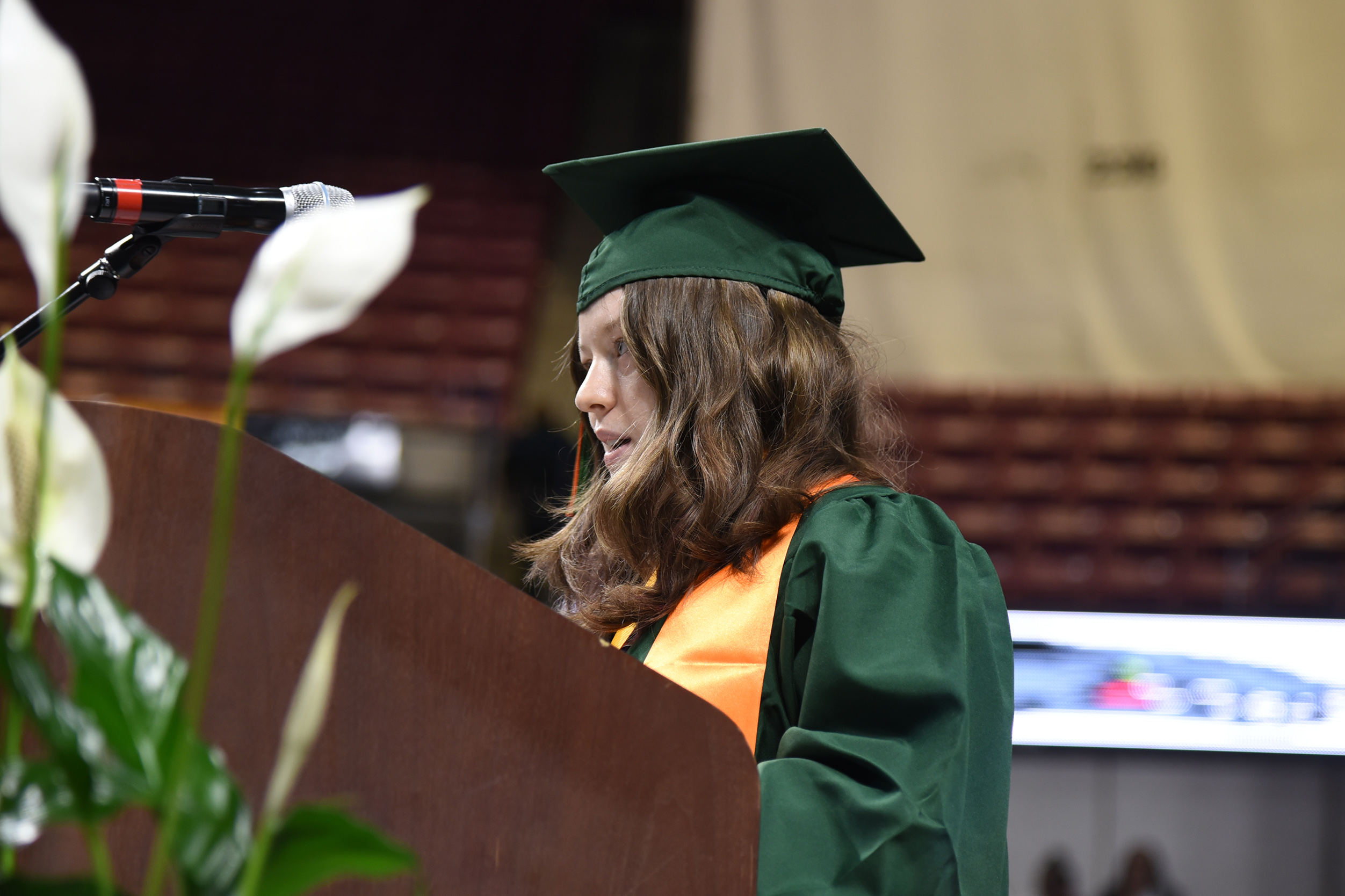 A woman in a green and orange graduation gown and cap is speaking at a podium.
