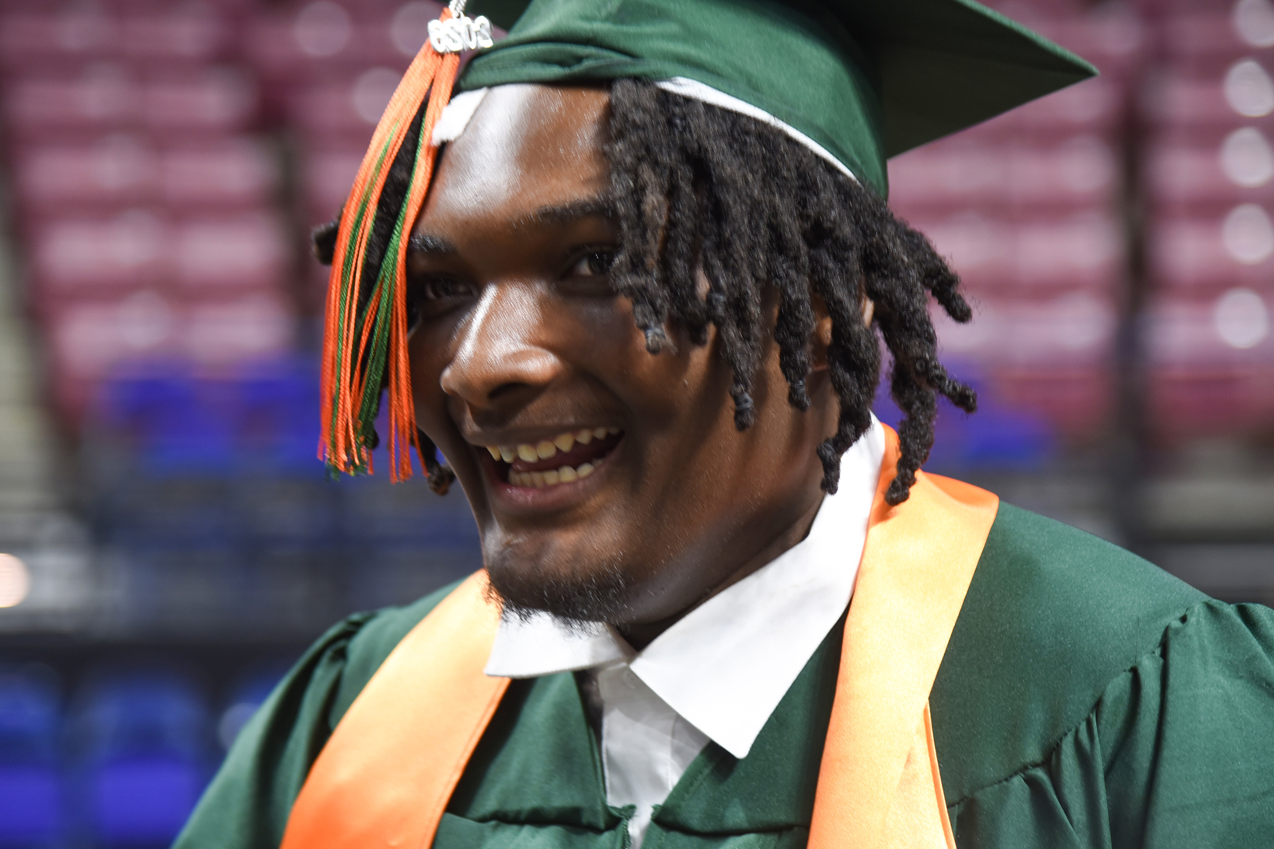 A man in a green cap and gown, smiling, stands against a blurred stadium background.