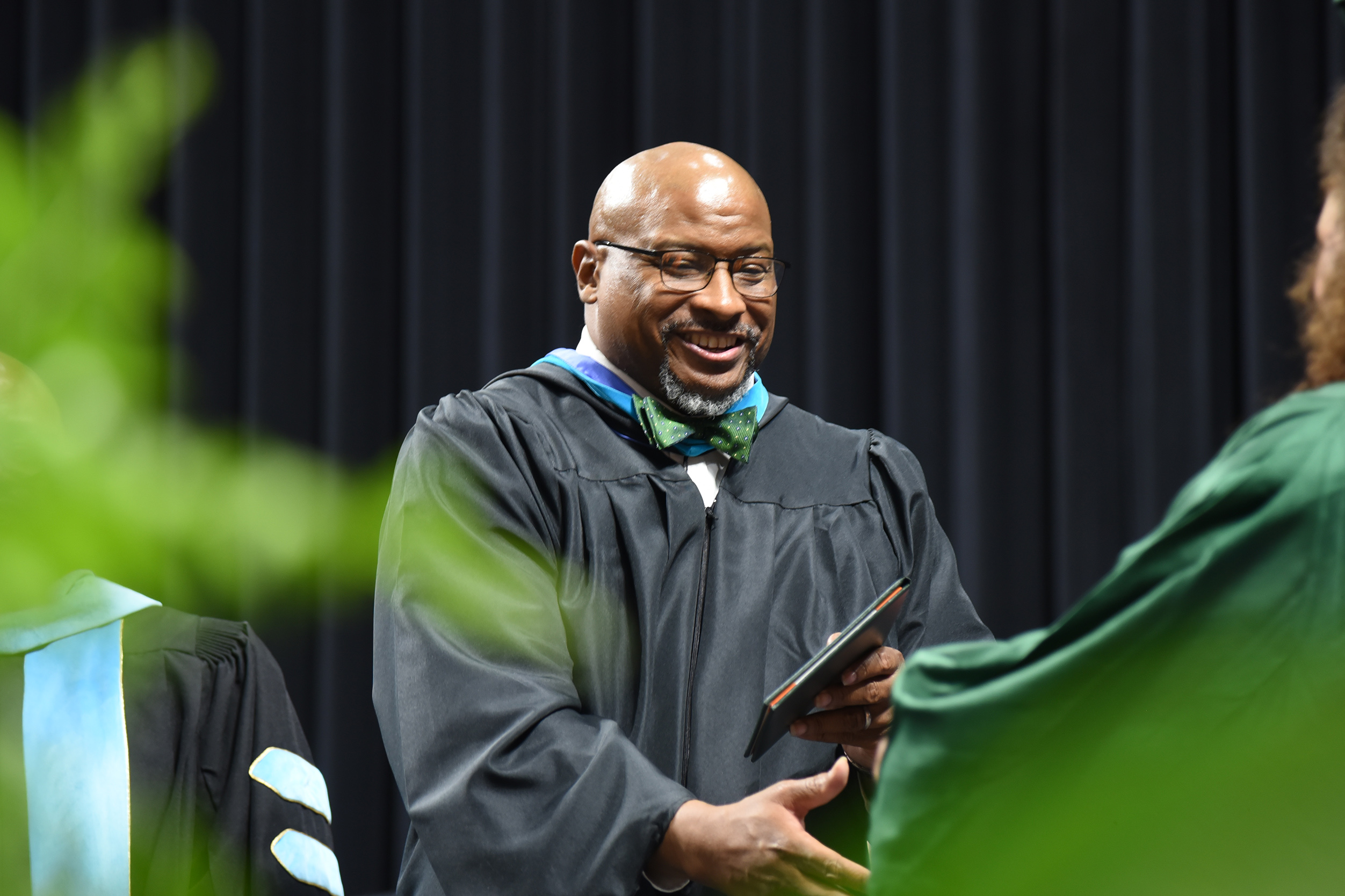 A man in a black graduation gown hands a diploma to a graduate in a green gown.