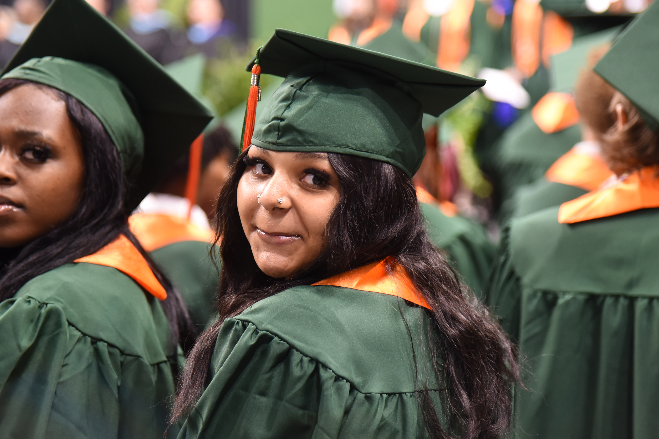 Graduates in green gowns and caps, one looking forward and smiling, in a crowded hall.