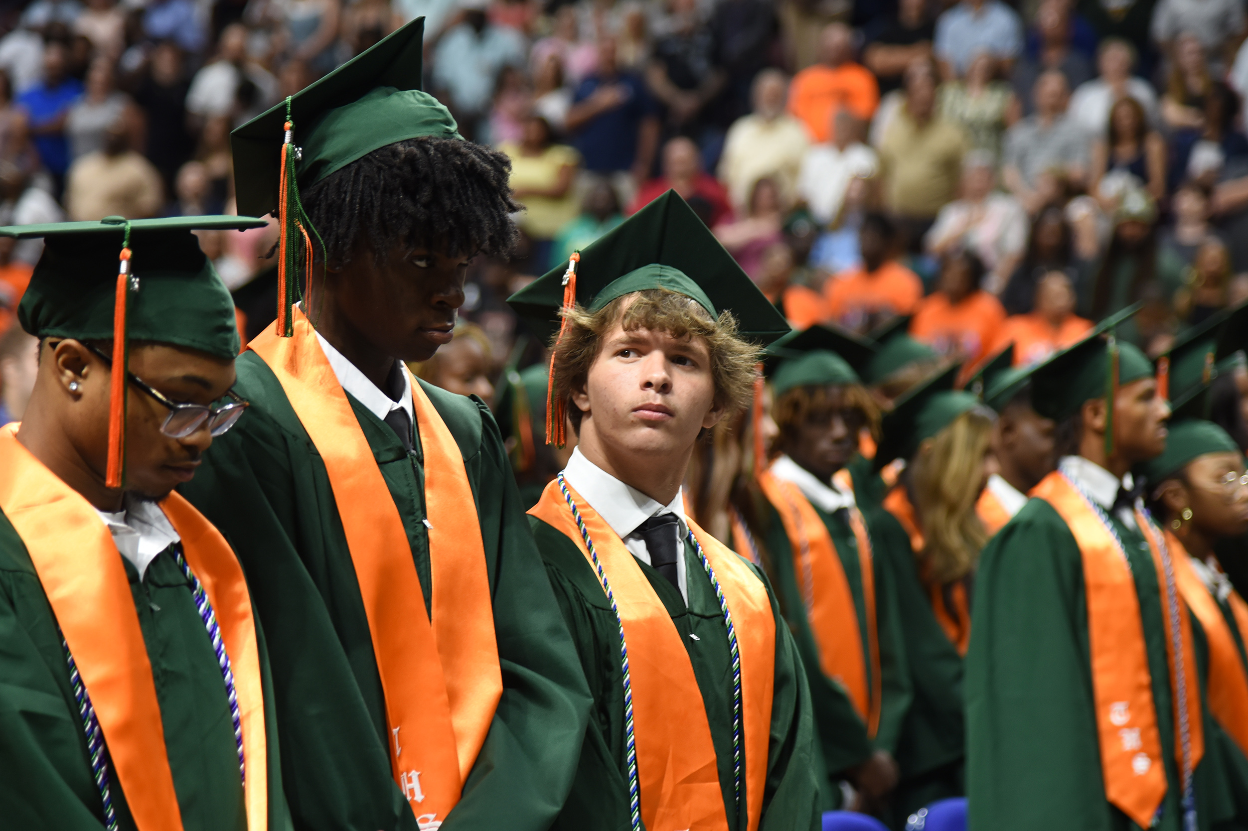 Graduating students in green and orange gowns, caps, and ties stand in line with audience behind.
