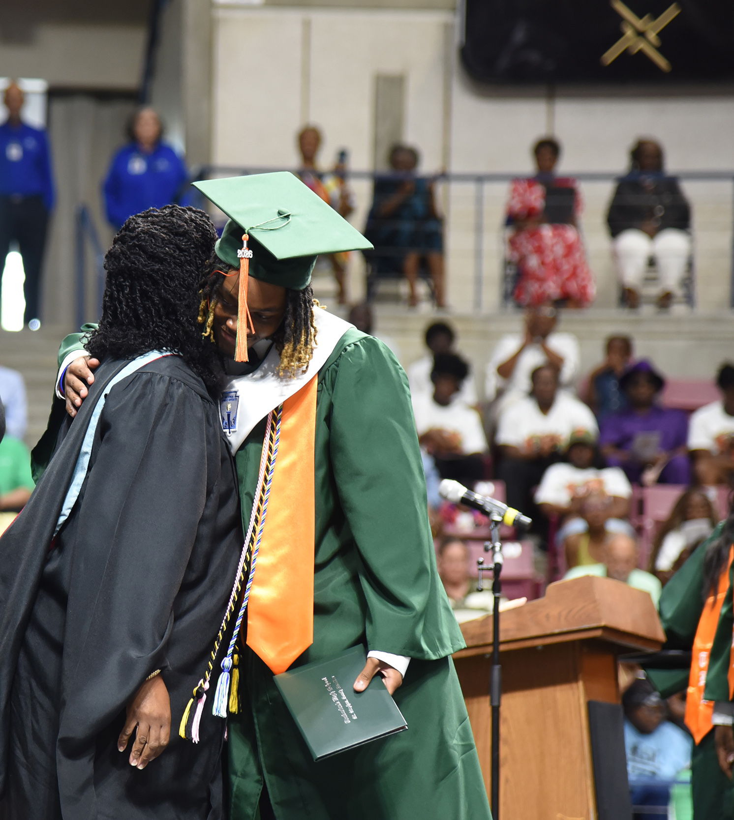 A graduate in a green gown and cap hugs a person in a black gown. Behind them, several people sit in rows.