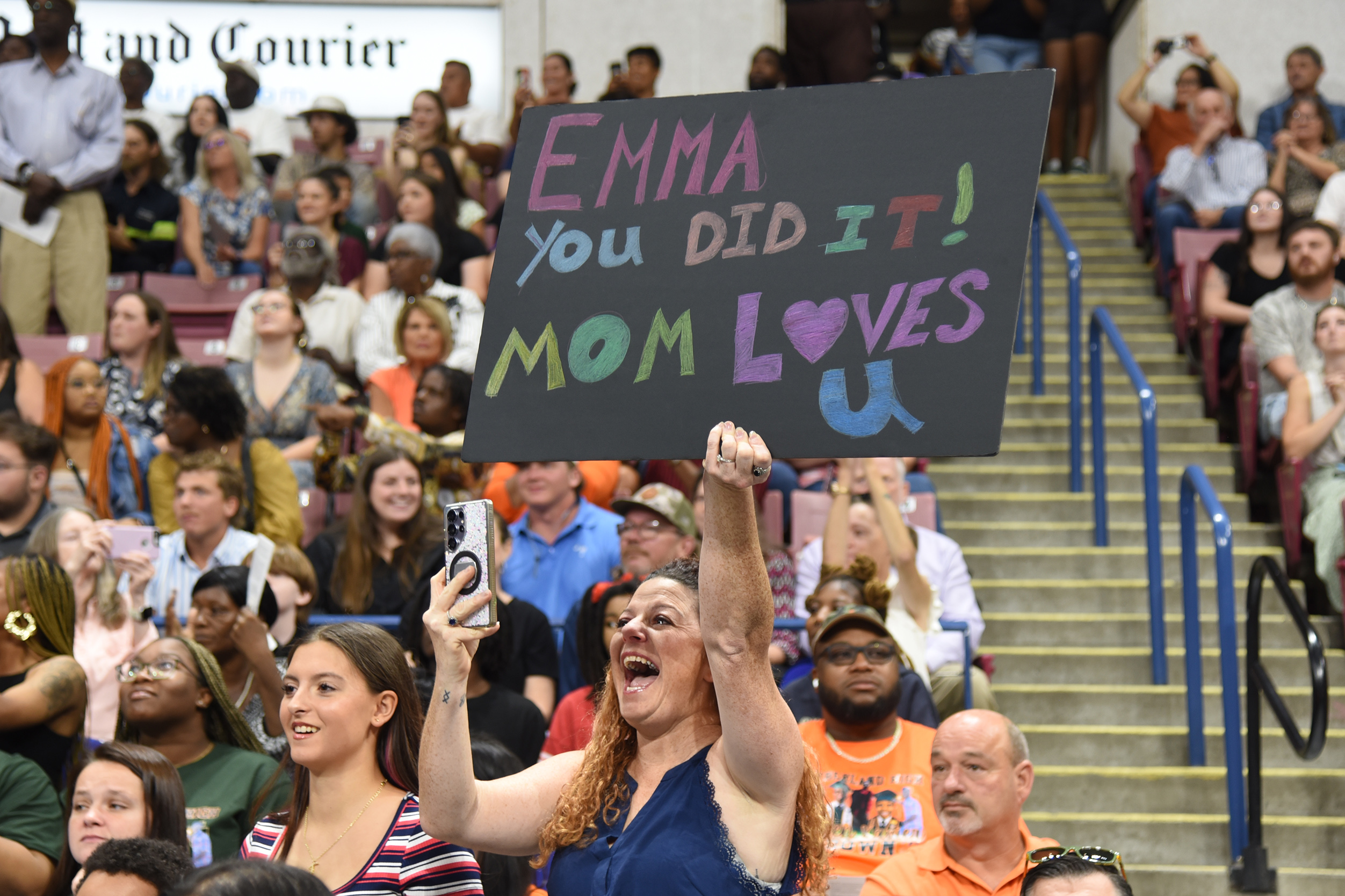 A crowd in a stadium, one person holds up a sign that says "Emma you did it! Mom loves you."