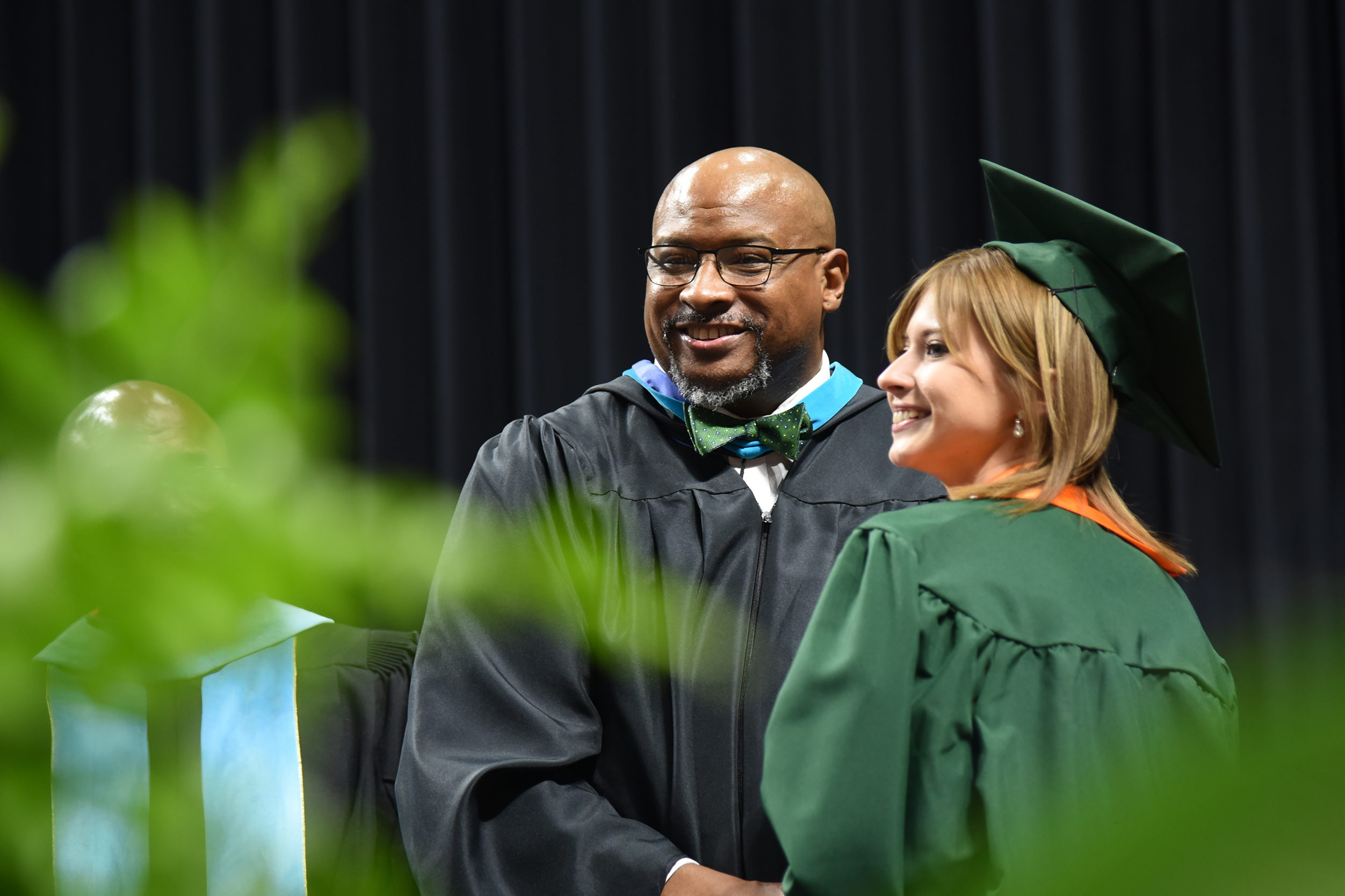 A man in a black graduation robe and glasses stands next to a woman in a green gown, both smiling.
