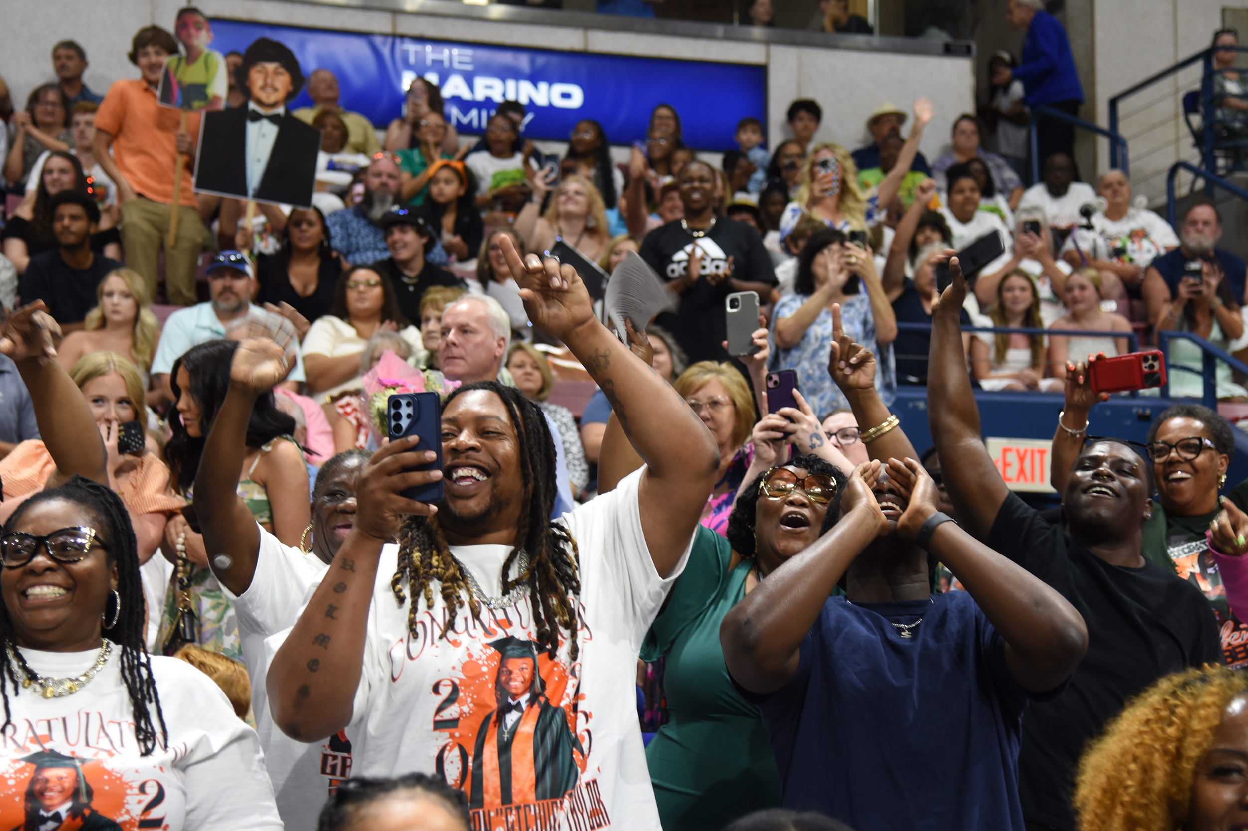 A crowd of people in a stadium, some raising hands, holding phones, and clapping. A blue banner with text is visible above.