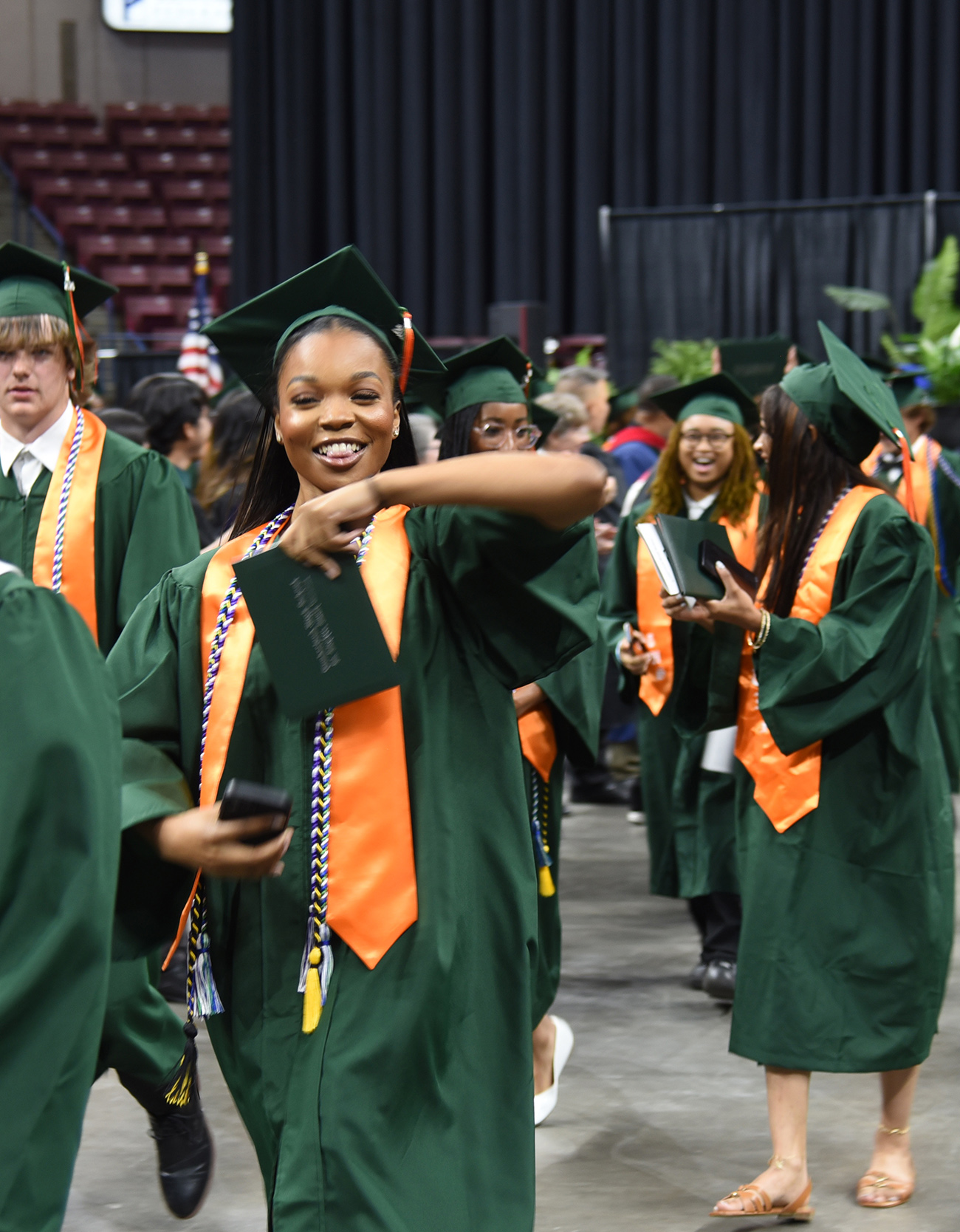 Graduates in green gowns and orange sashes walk in an indoor ceremony. One woman holds a book and smiles.