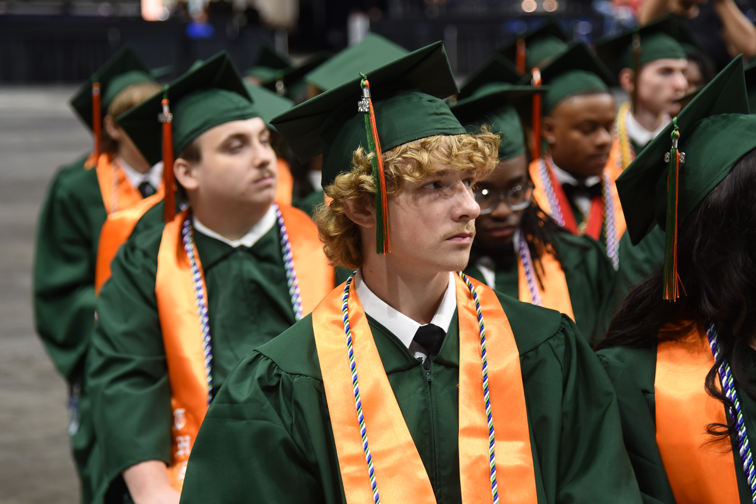 A group of graduates in green caps and gowns, some with orange sashes, line up for a ceremony.