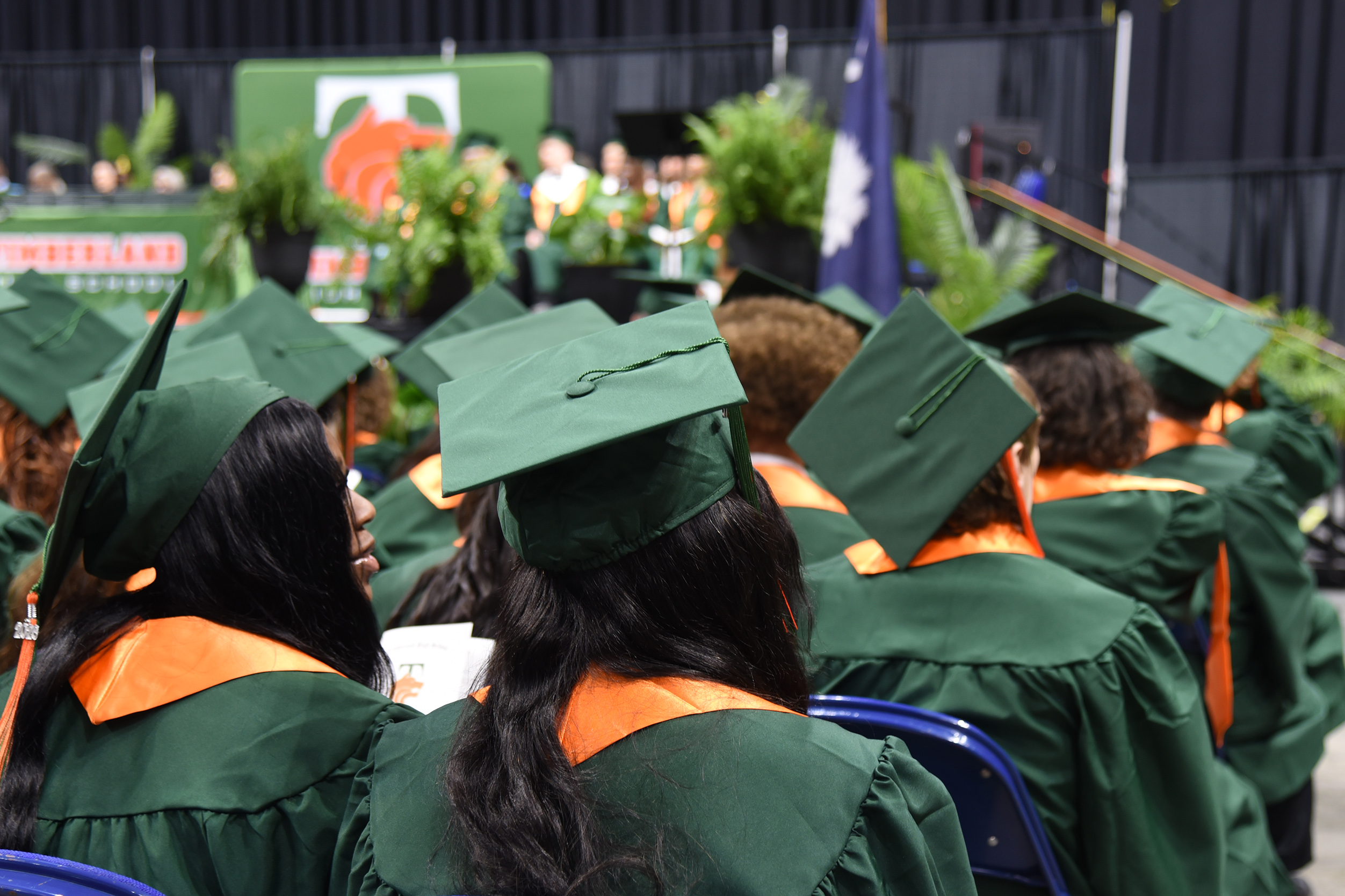 Group of graduates in green caps and gowns at a ceremony. Some are looking forward; others are facing sideways.