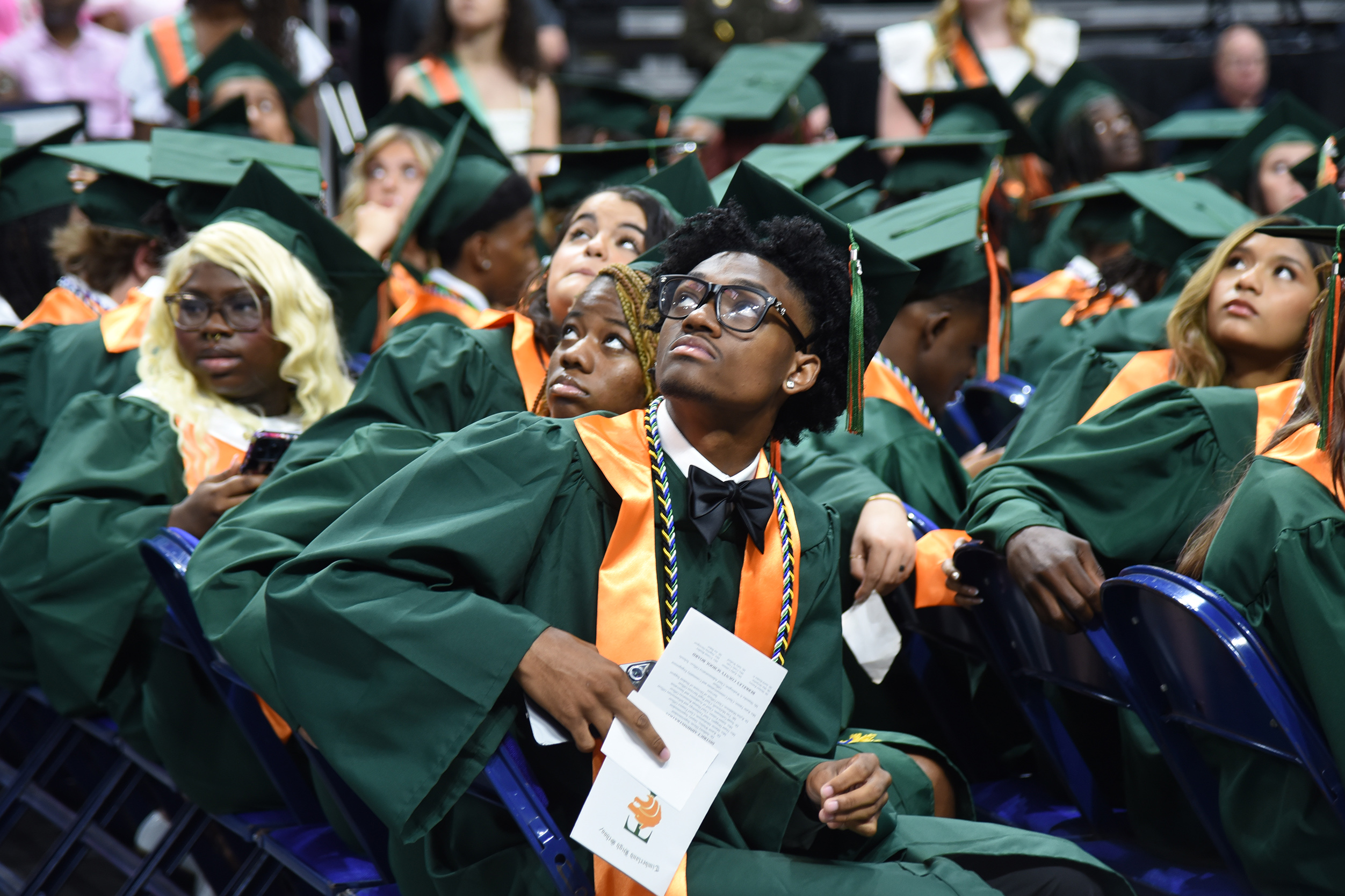 A group of people in green gowns and caps sitting in chairs at a graduation ceremony.