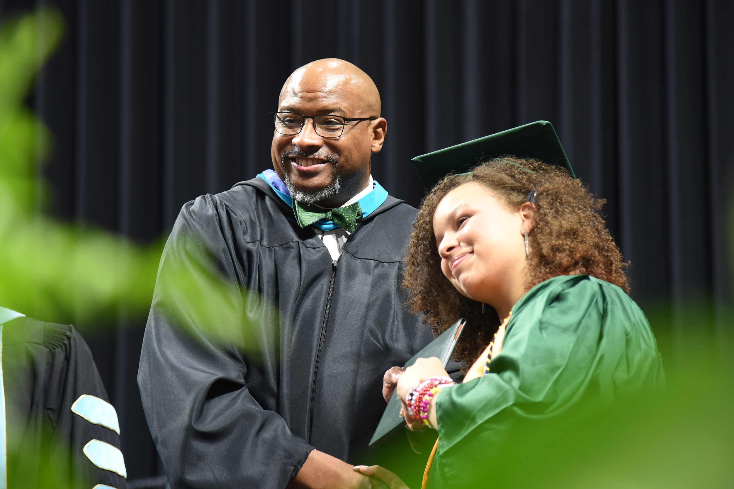 A man and woman in academic robes. The woman in green holds a diploma. The man smiles.
