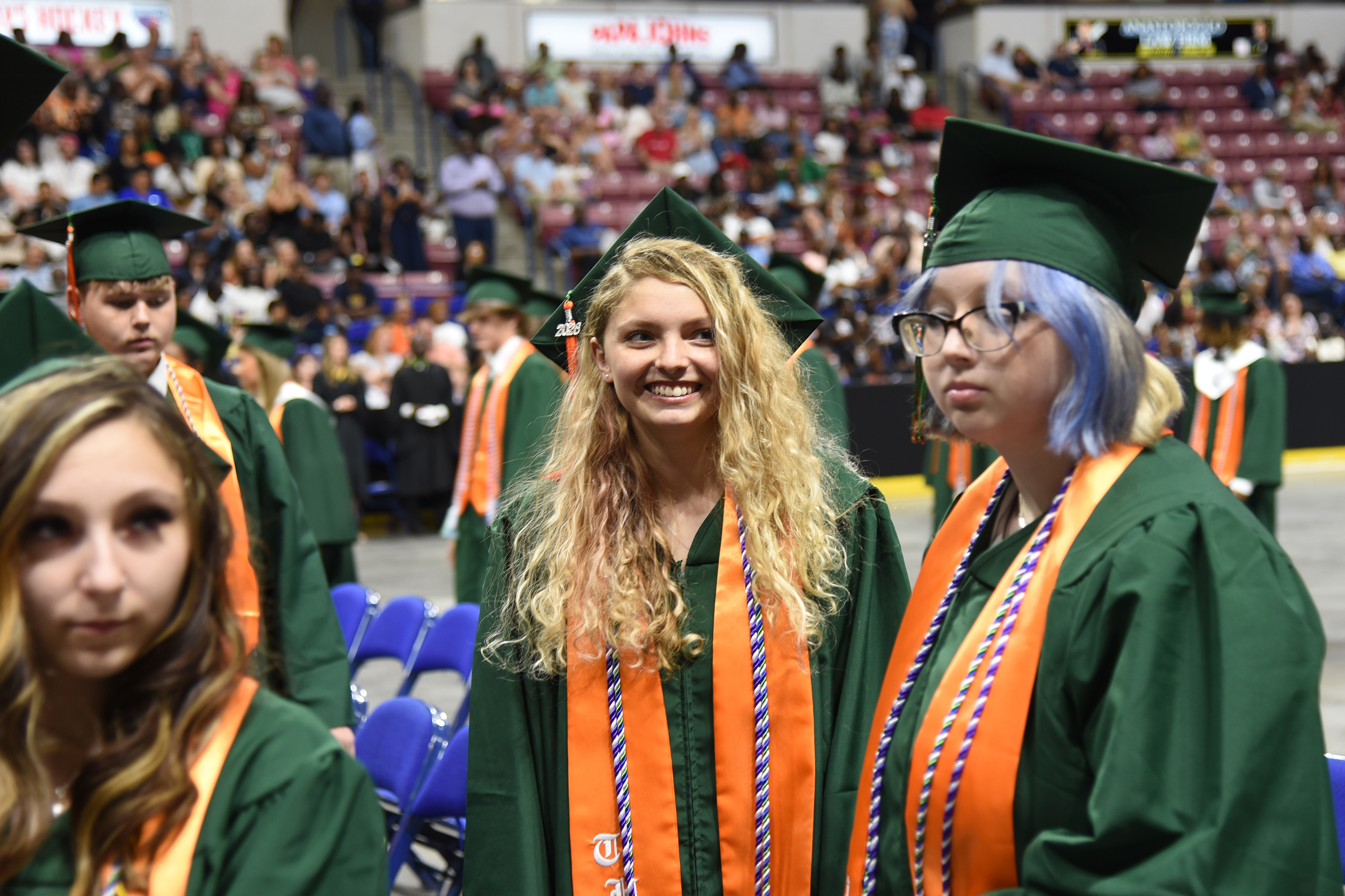 Three women in green gowns and mortarboard caps stand in a line at a graduation ceremony.