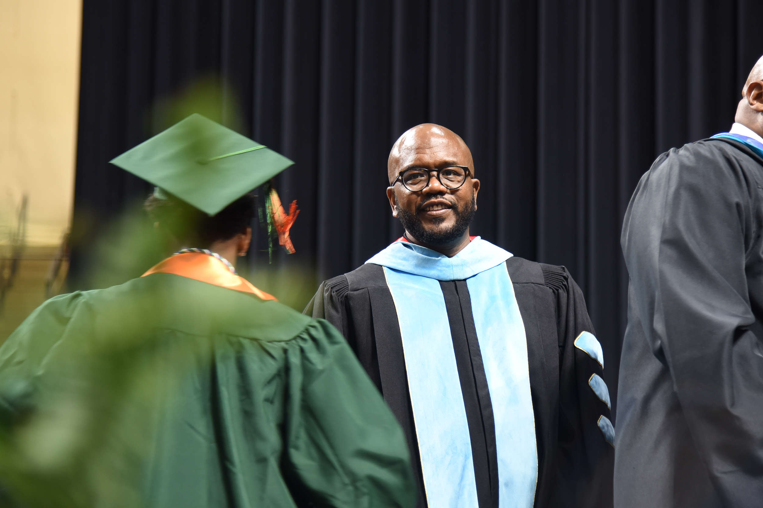 A man in academic regalia stands beside another graduate wearing a green cap and gown. Black curtains hang in the background.