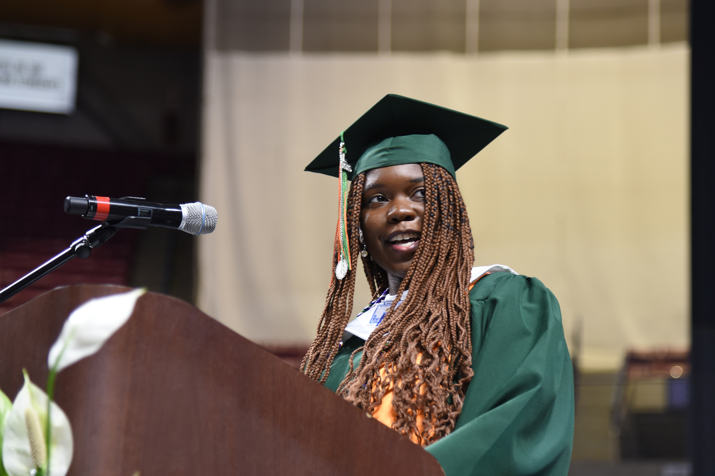 A young woman with a green gown and cap stands at a podium with a microphone.