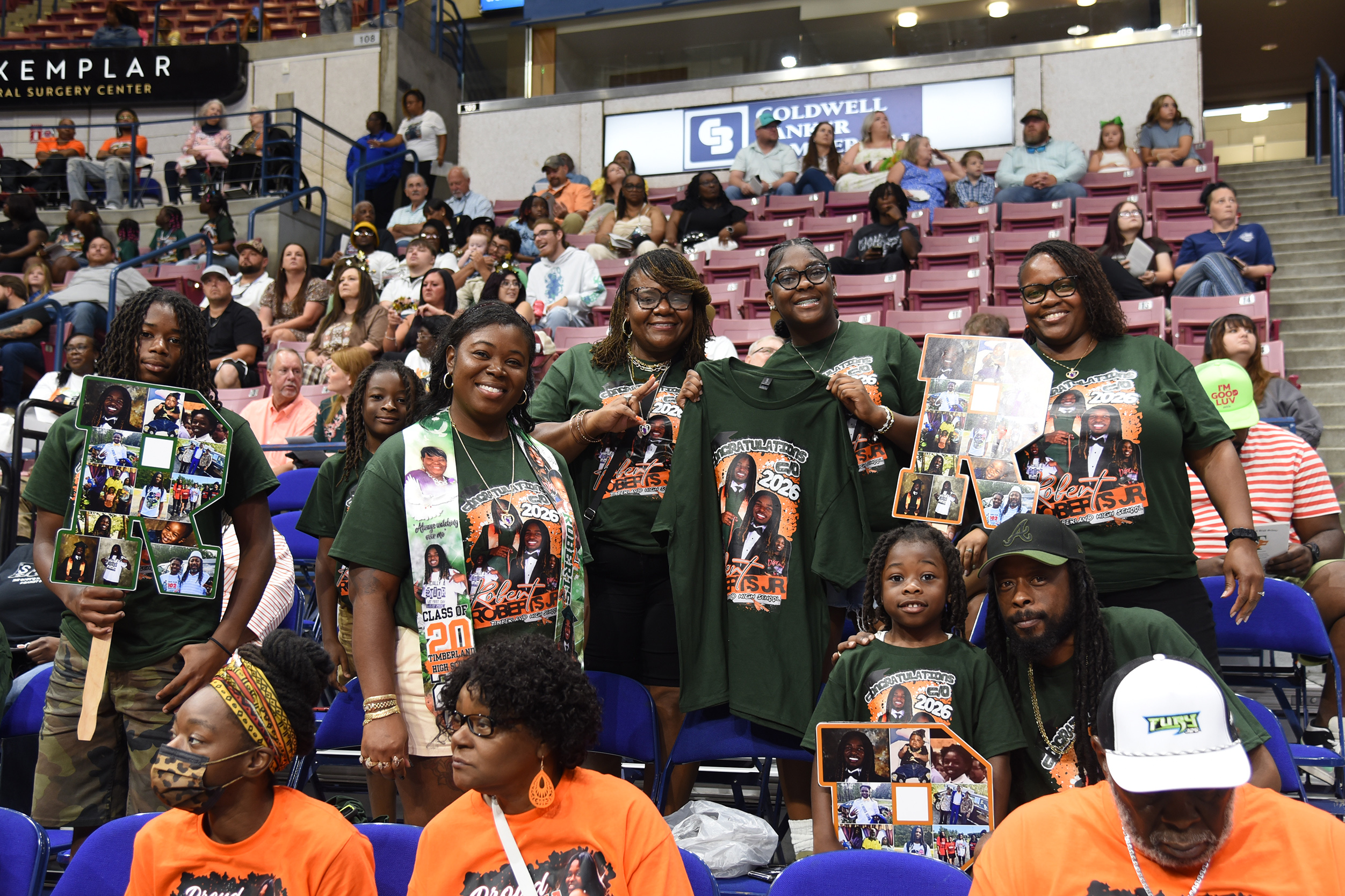Group of people in matching shirts holding signs with photos, seated in a stadium with bleacher seating.