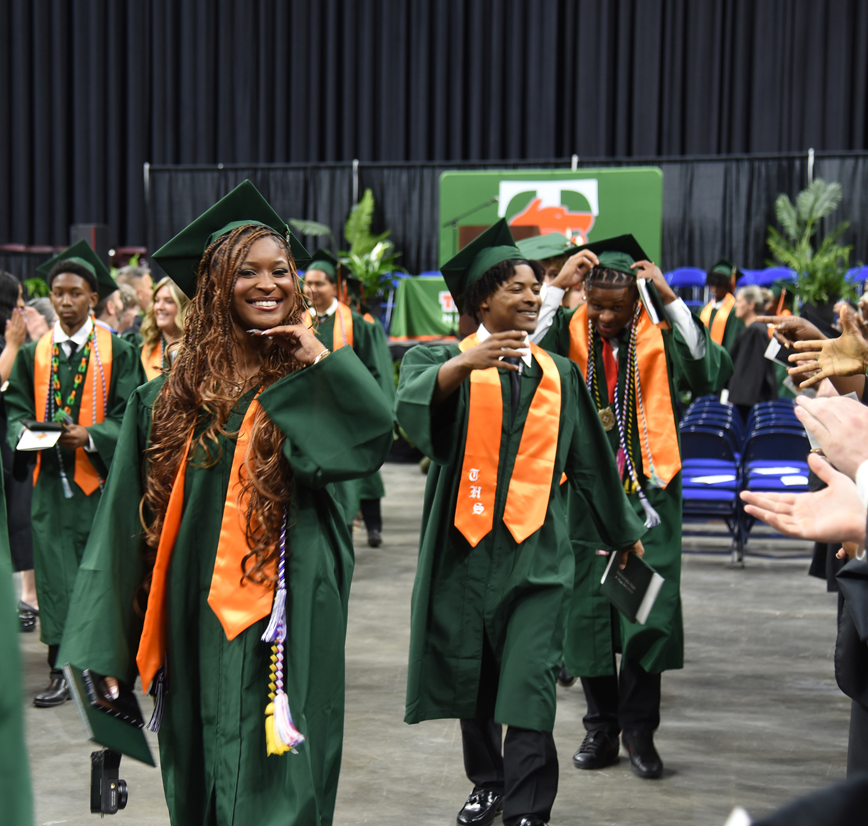 People in green caps and gowns walk past clapping hands. Some wear orange sashes. The background has greenery and a green banner.