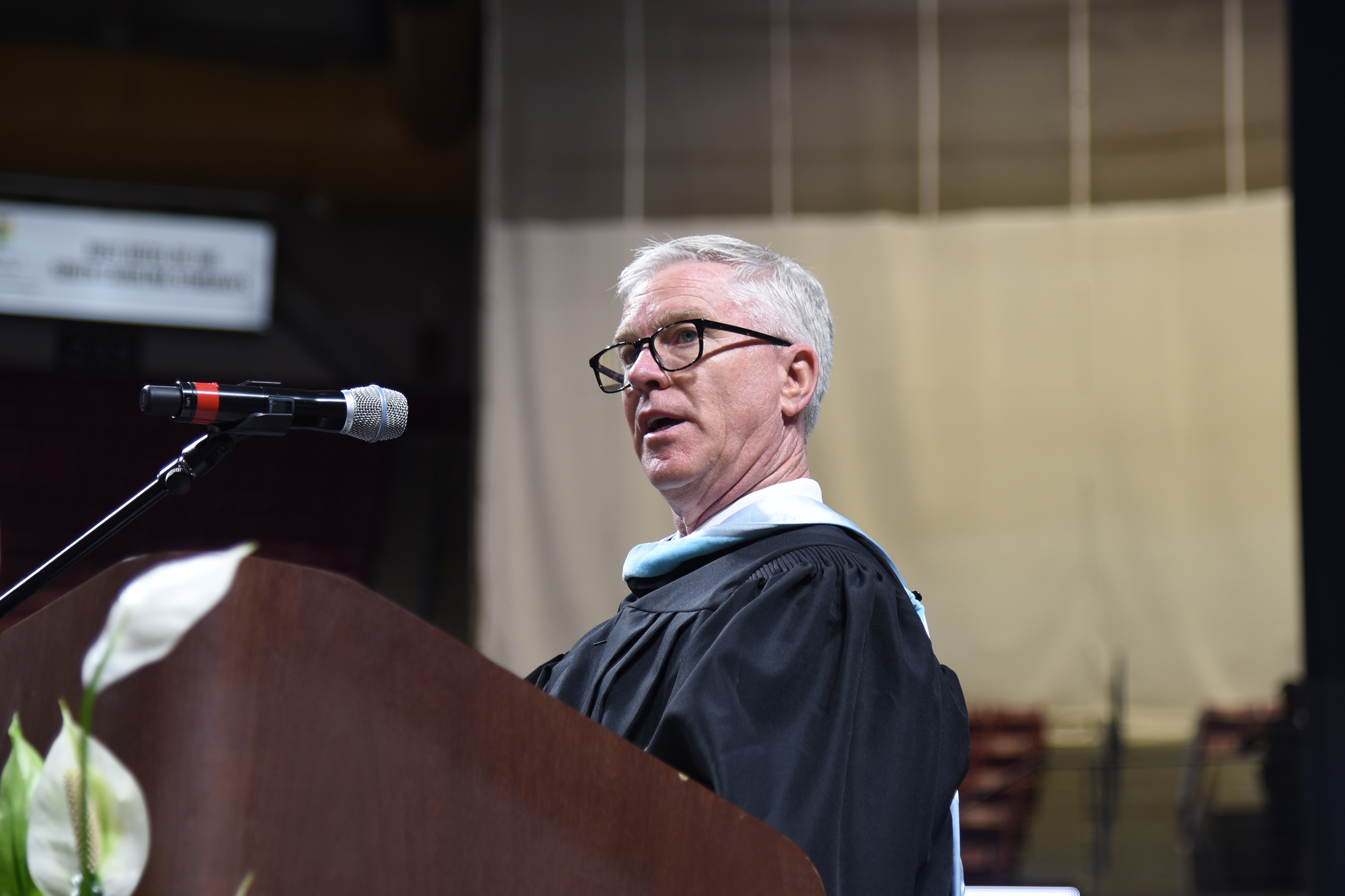 A man in a black gown and glasses speaks at a podium with a microphone.