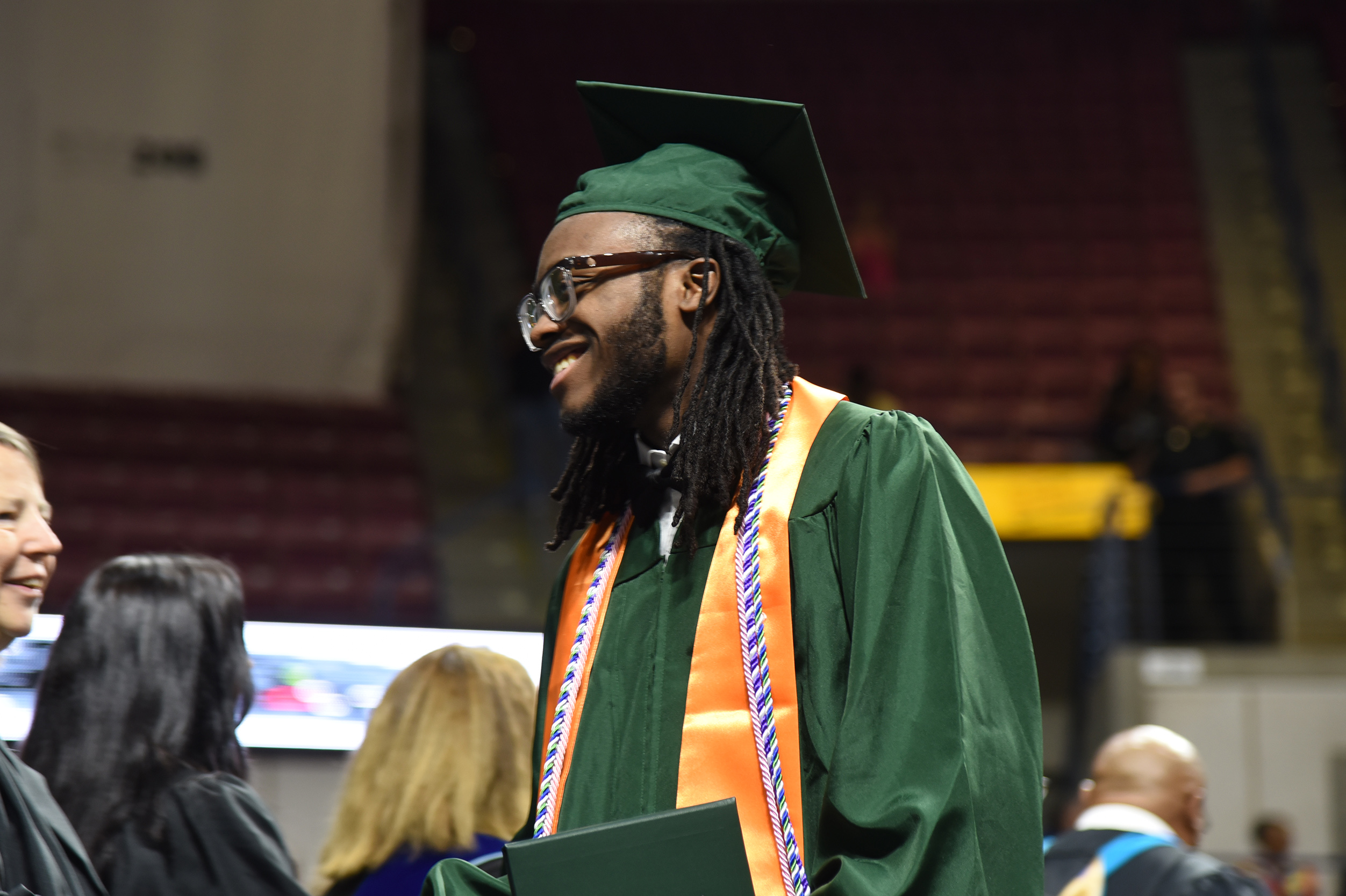 A man in a green graduation gown and hat stands in a stadium. Other people stand behind him.