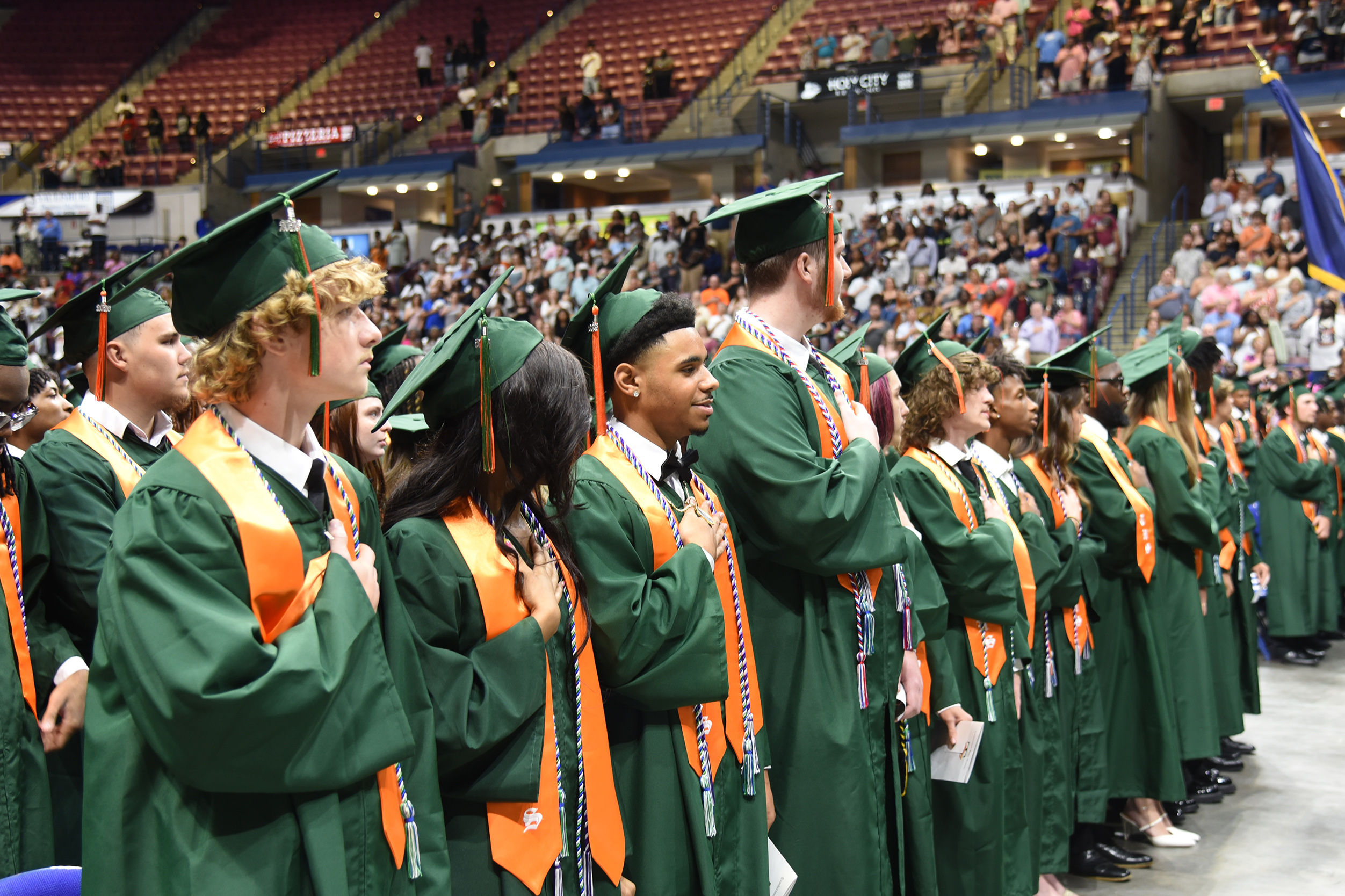 Graduates wearing green caps and gowns stand in a line, facing a large audience in an indoor stadium.
