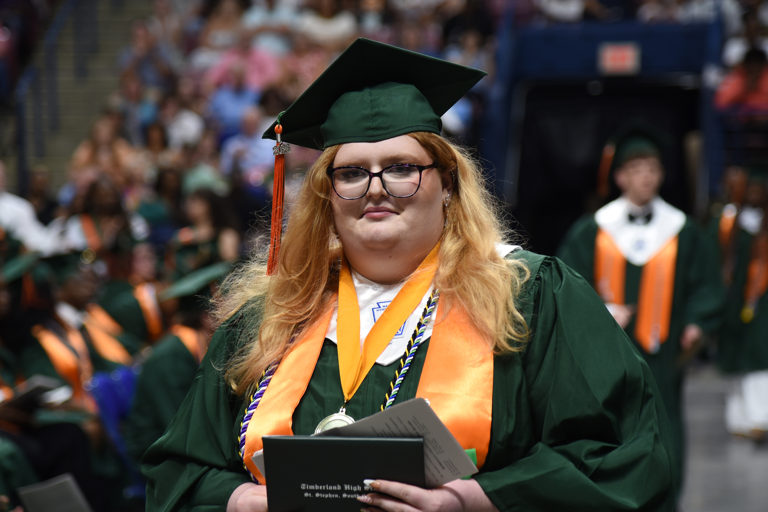 Person with a graduation cap and gown stands in a crowd holding a diploma and certificate.