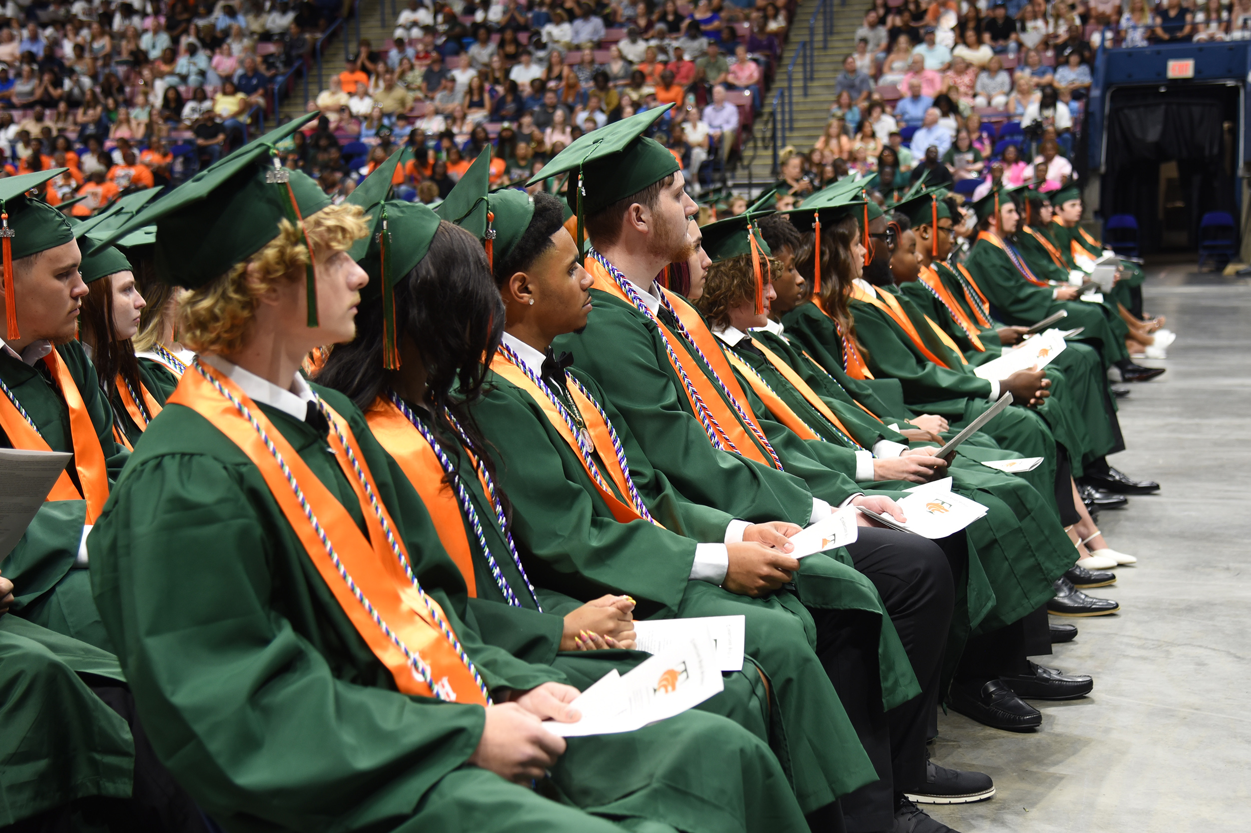Graduates in green gowns and mortarboards sit in rows, holding diplomas, with a large audience in the background.