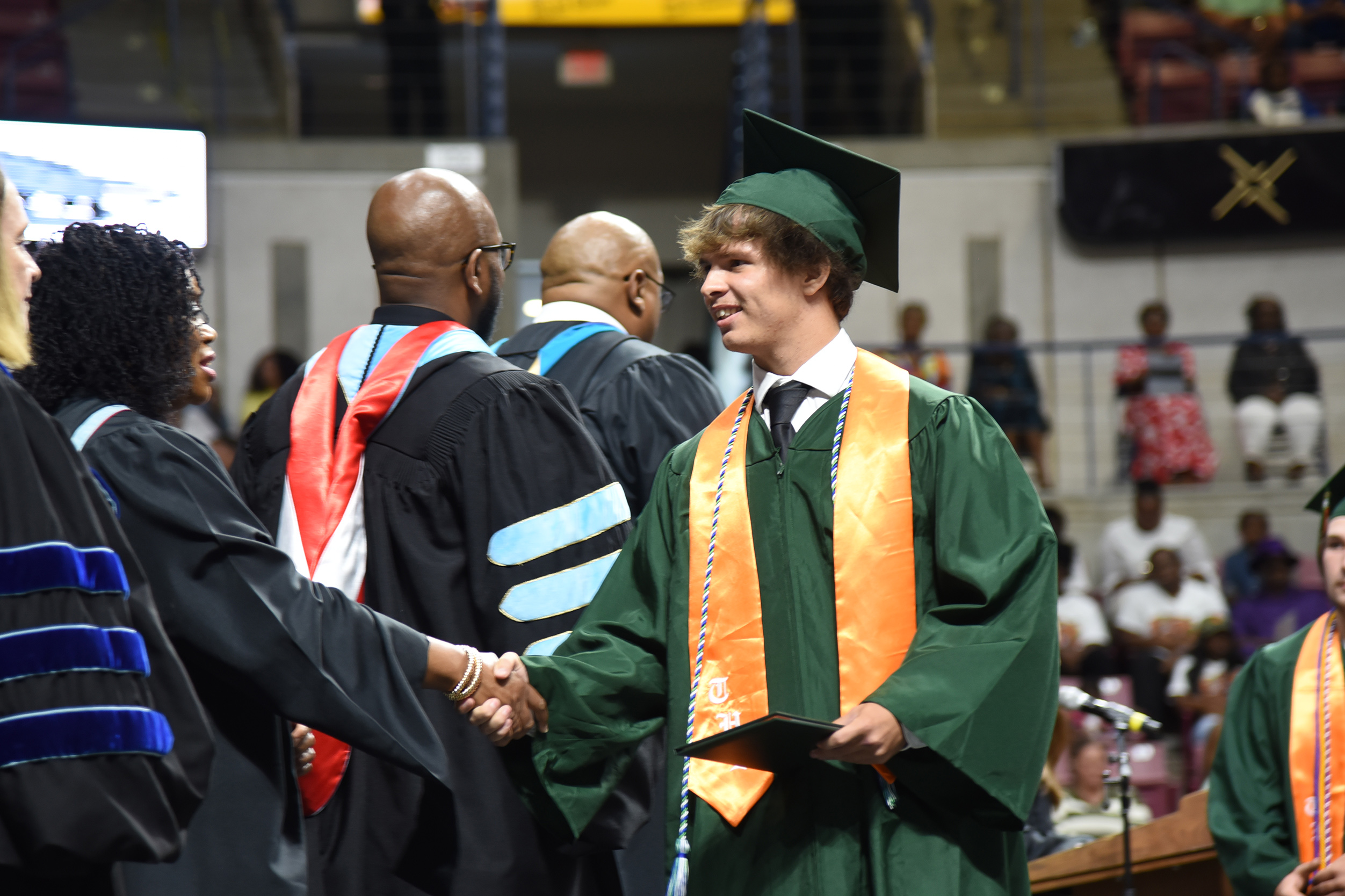 A man in a green graduation gown and cap shakes hands with another individual at a ceremony.