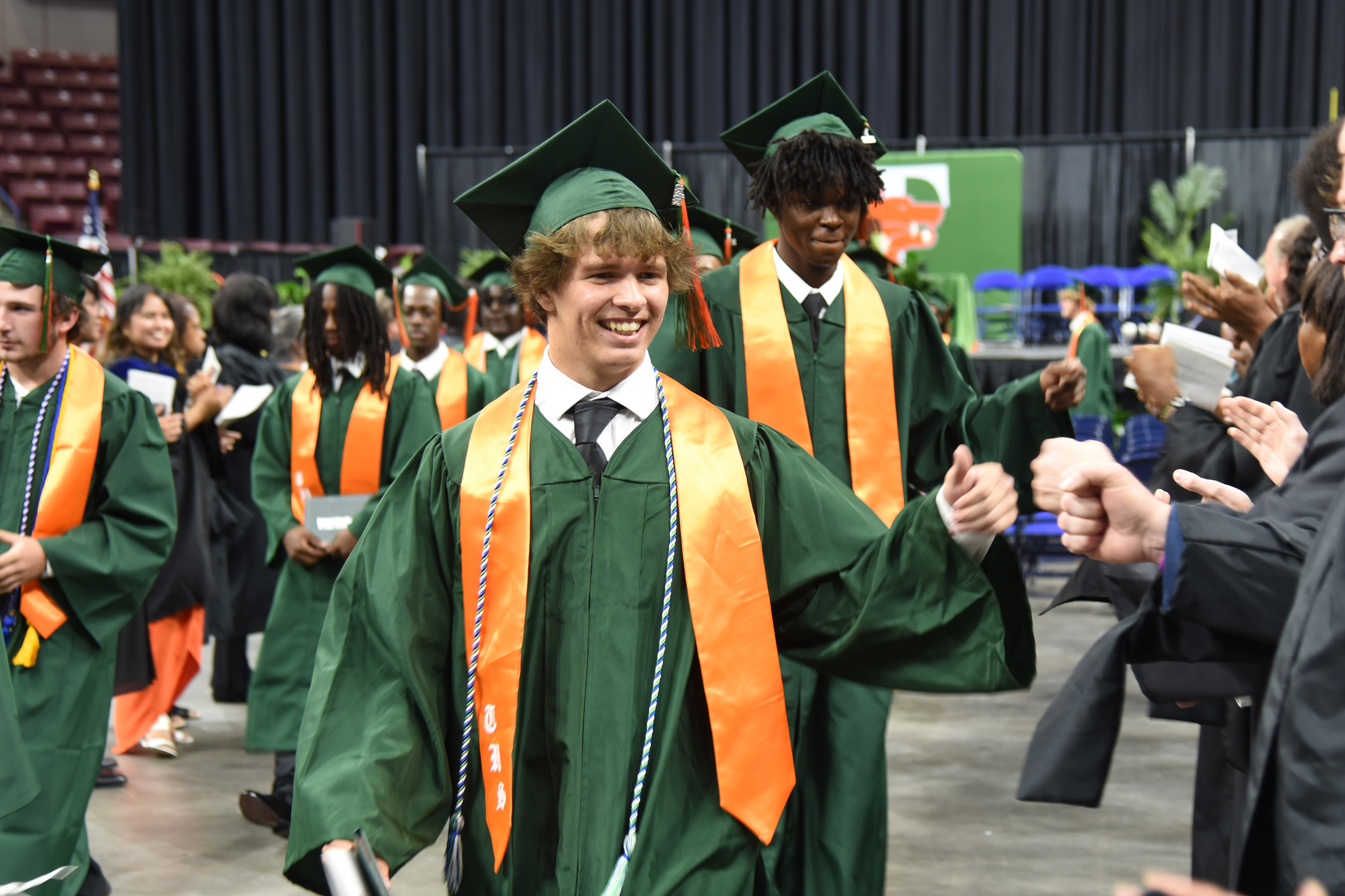 Graduates in green gowns walk toward the audience, one smiling. The background shows a large open space.
