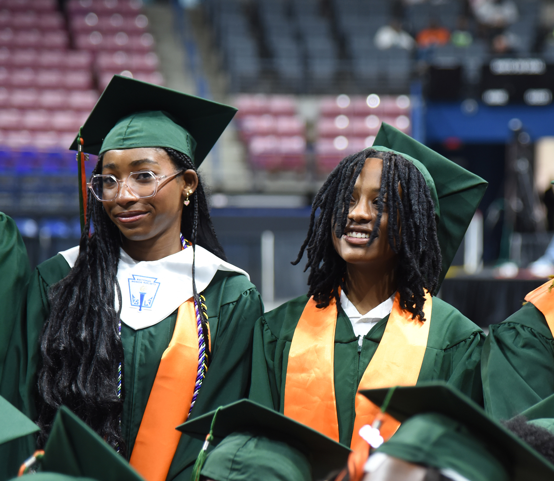 Two women in green graduation gowns and caps stand among others in a stadium with red seats.
