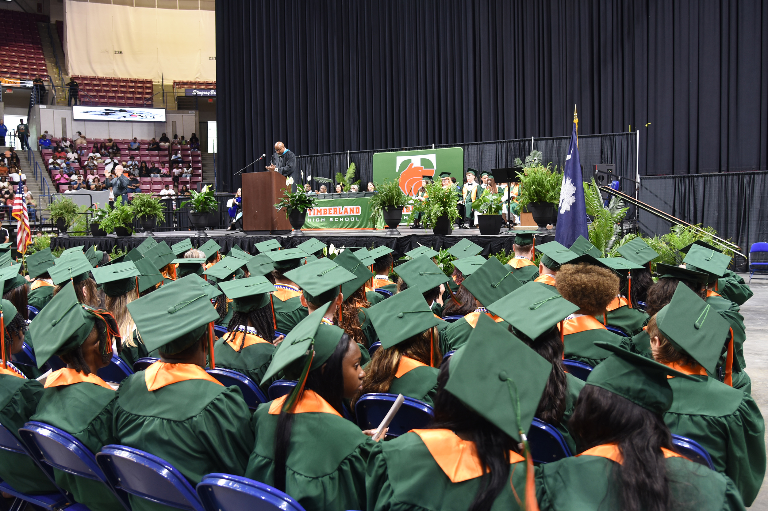A crowd of graduates in green gowns and caps seated in rows at an indoor graduation ceremony.