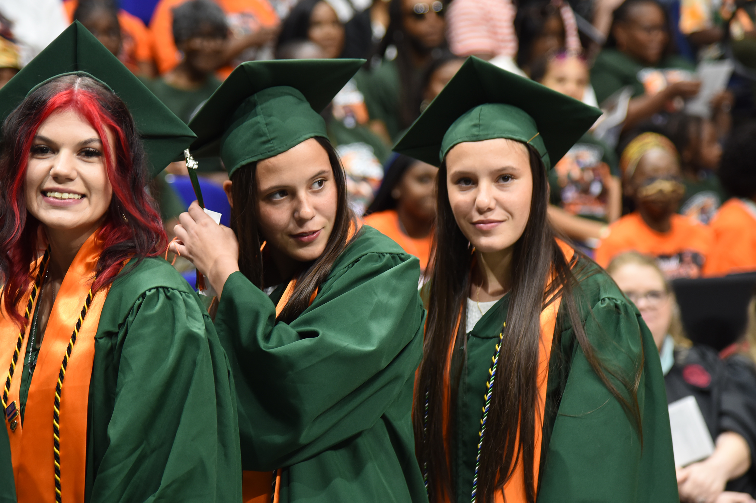 Three women in green graduation gowns with orange sashes smile, standing before a crowd.