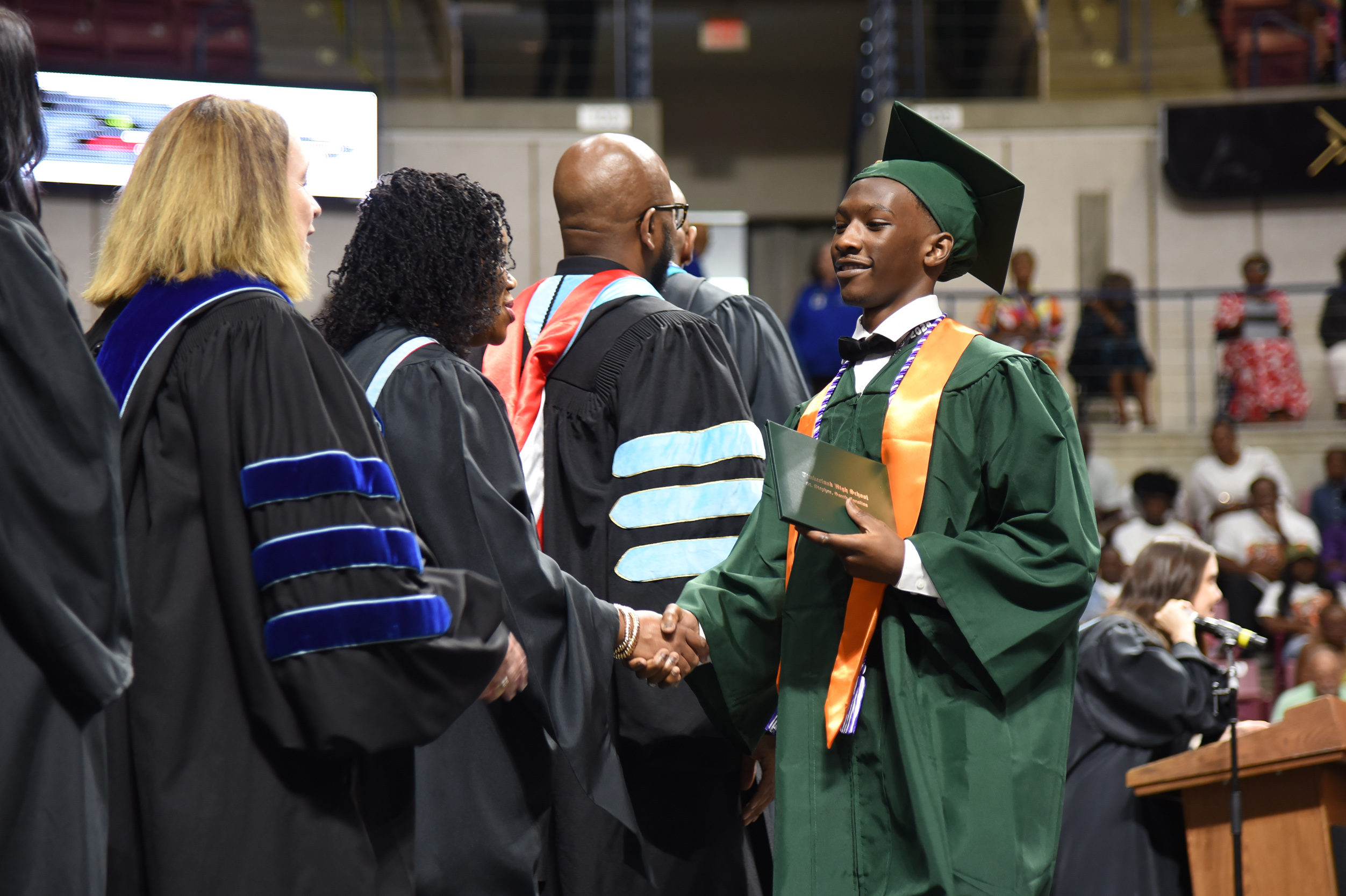 A young person in green and gold shakes hands with a woman in a black gown.