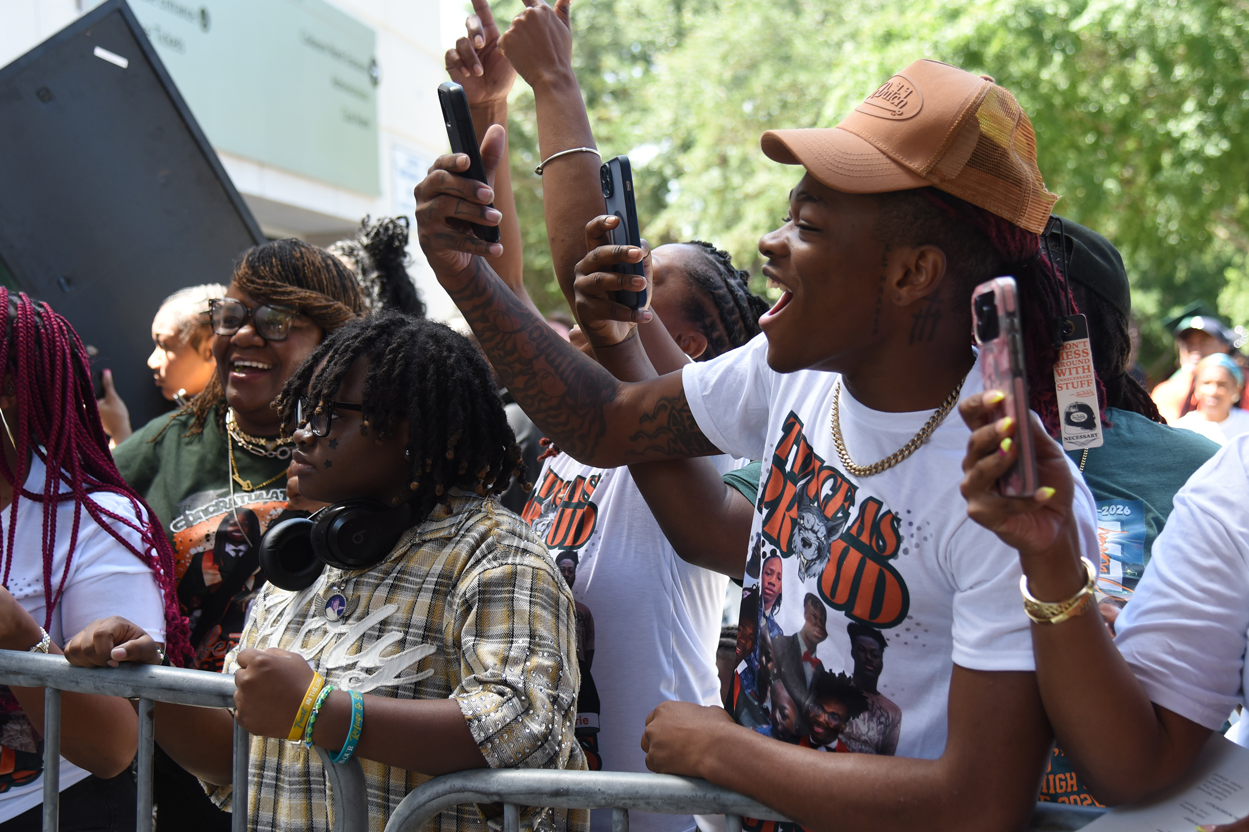 Group of people at an outdoor event, one wearing a cap, raising hands with cell phones.