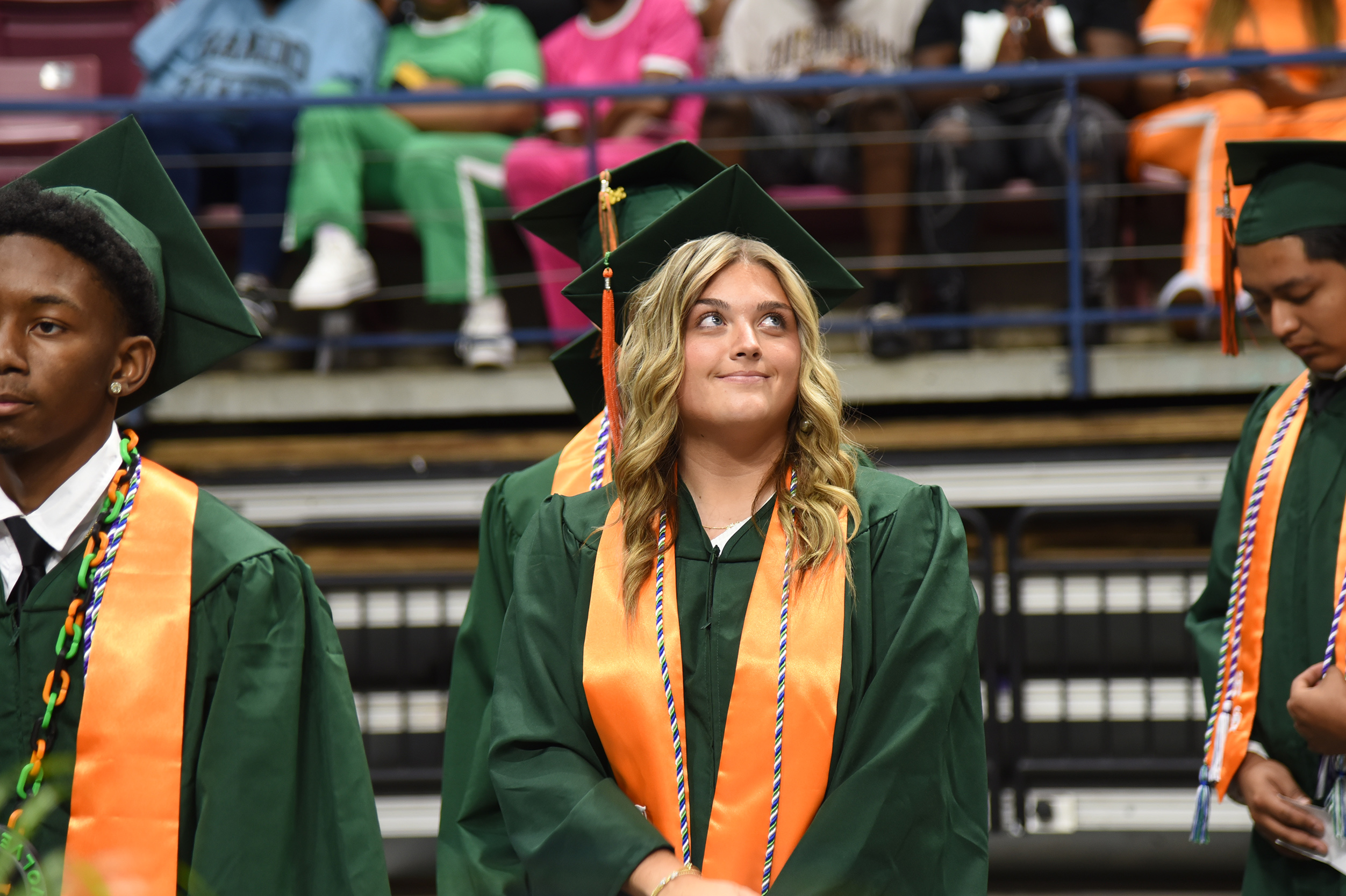 Four people in green graduation gowns with orange sashes stand before a bleachers section.