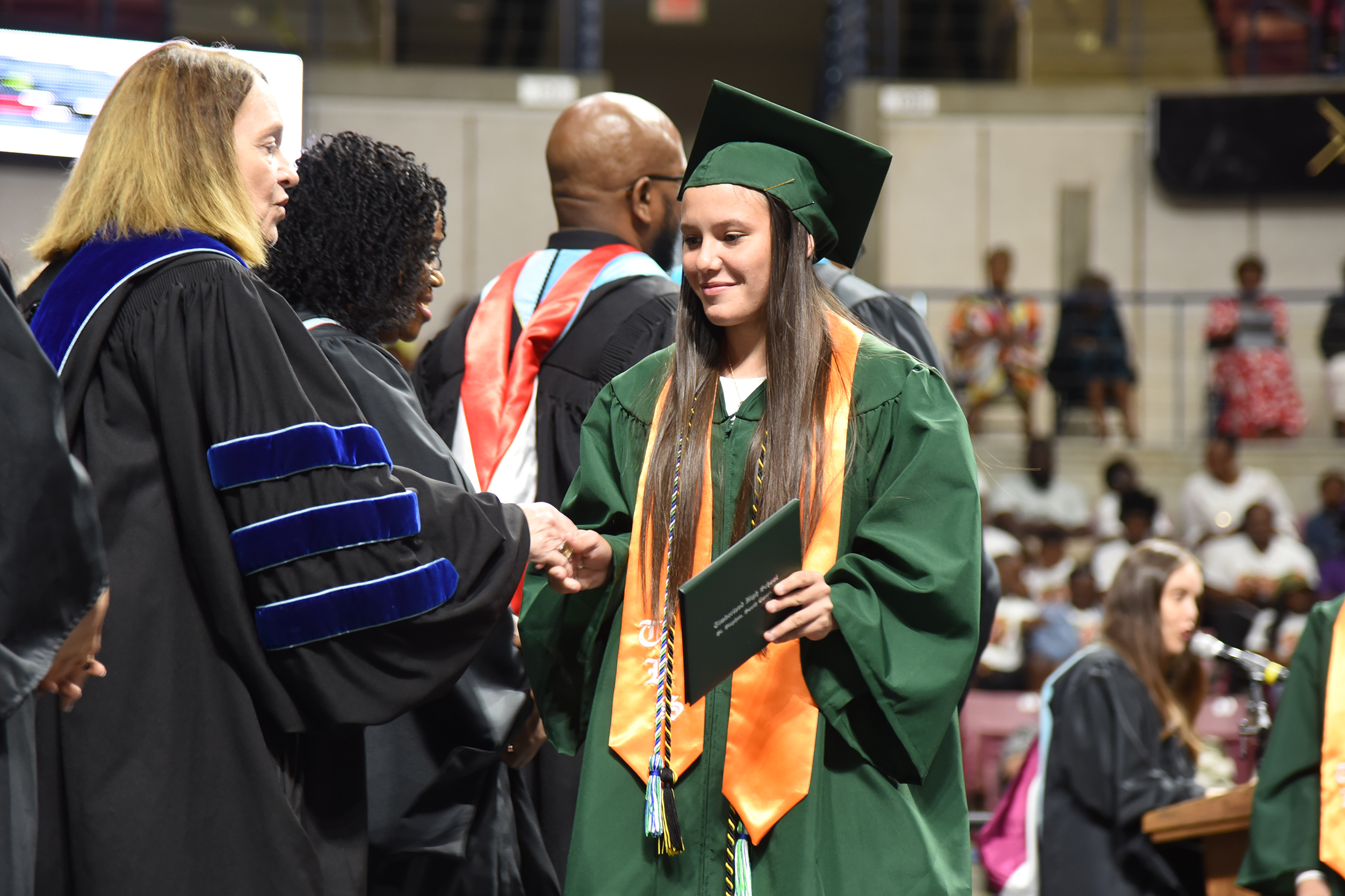 A woman in a green gown shakes hands with a faculty member in a black gown during a graduation ceremony.