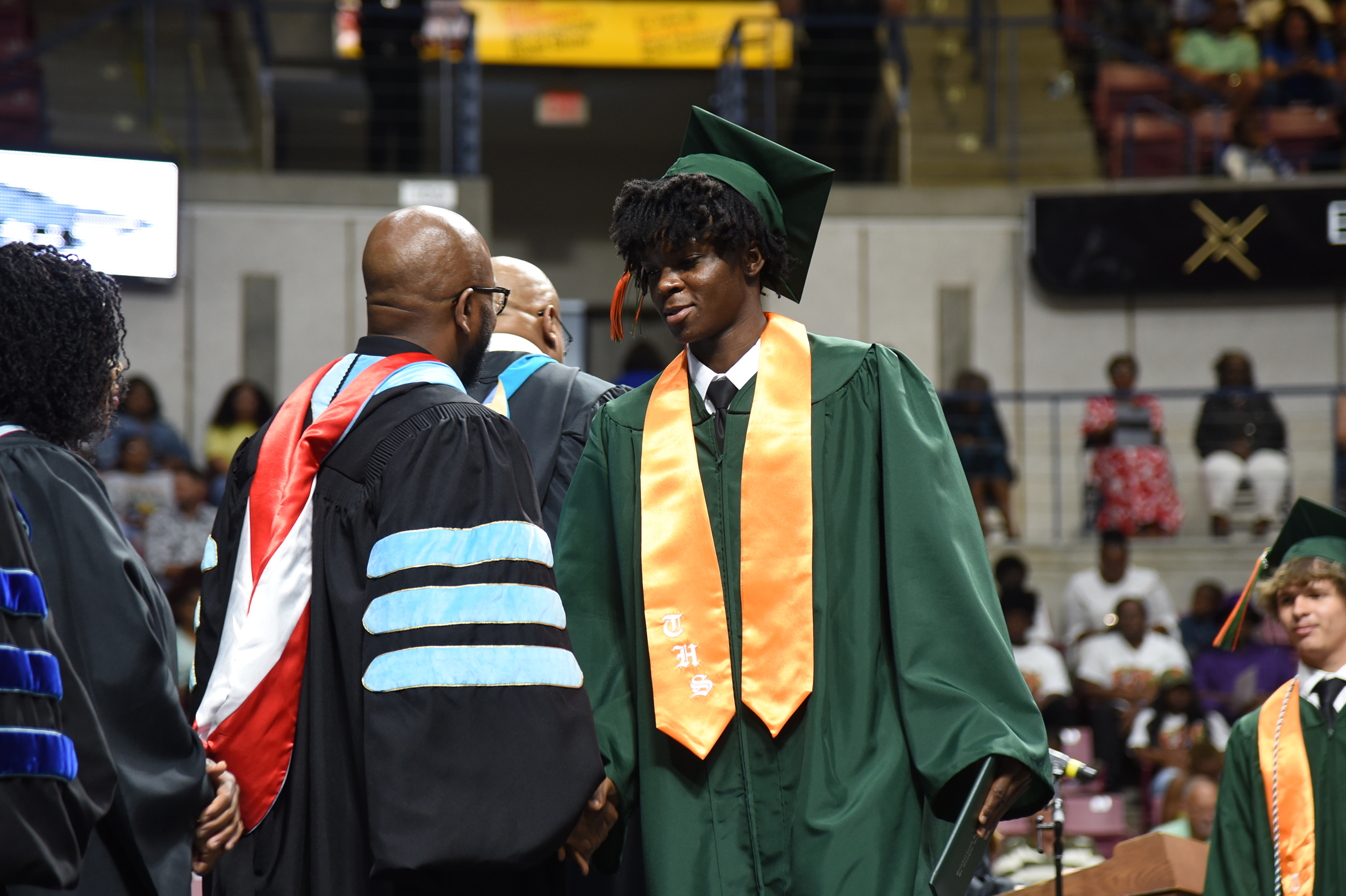 People at a graduation ceremony, including a student in green, orange, and black graduation garb.