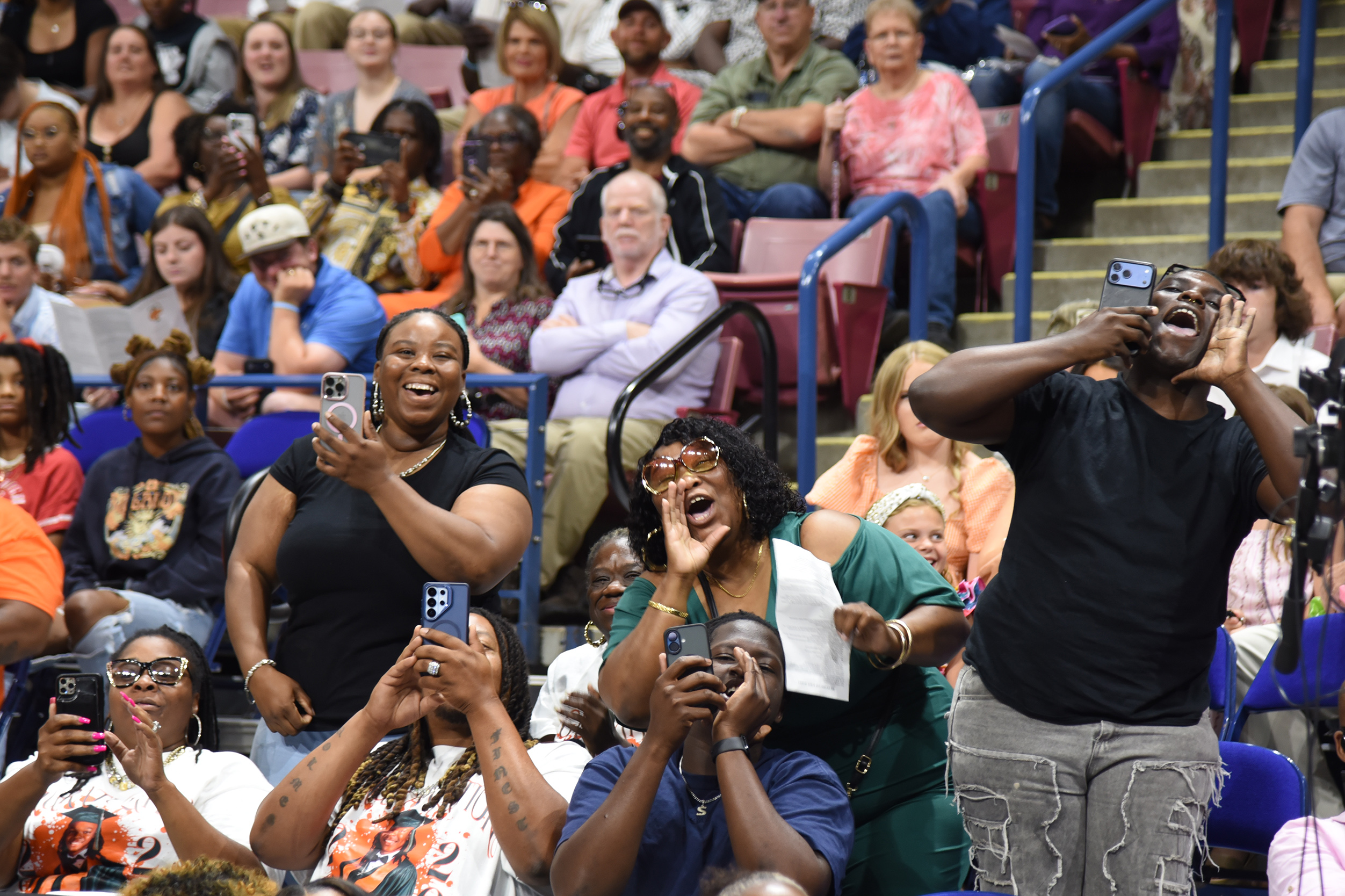 Spectators at a sporting event are cheering and recording with phones. Some wear glasses and bracelets.