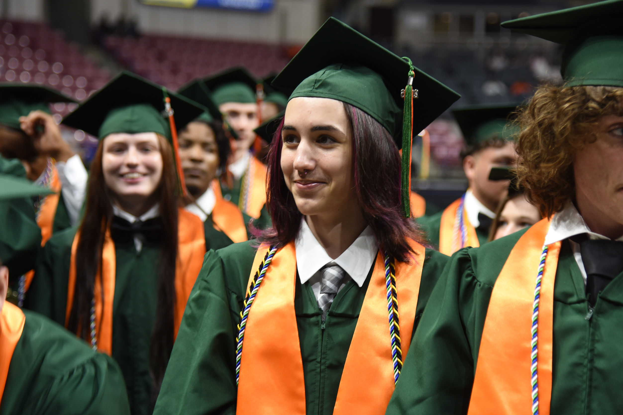 A group of students in green graduation gowns and orange sashes, possibly at a graduation ceremony.