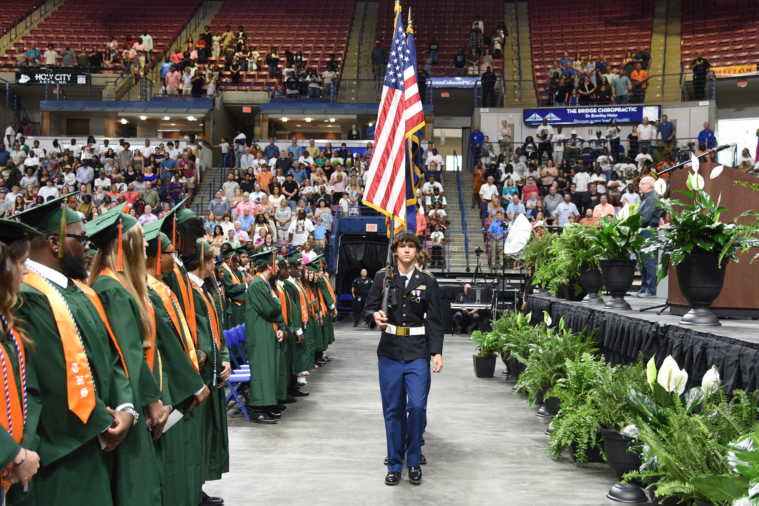 Military member walks between graduates during a graduation ceremony. Audience seated in bleachers.
