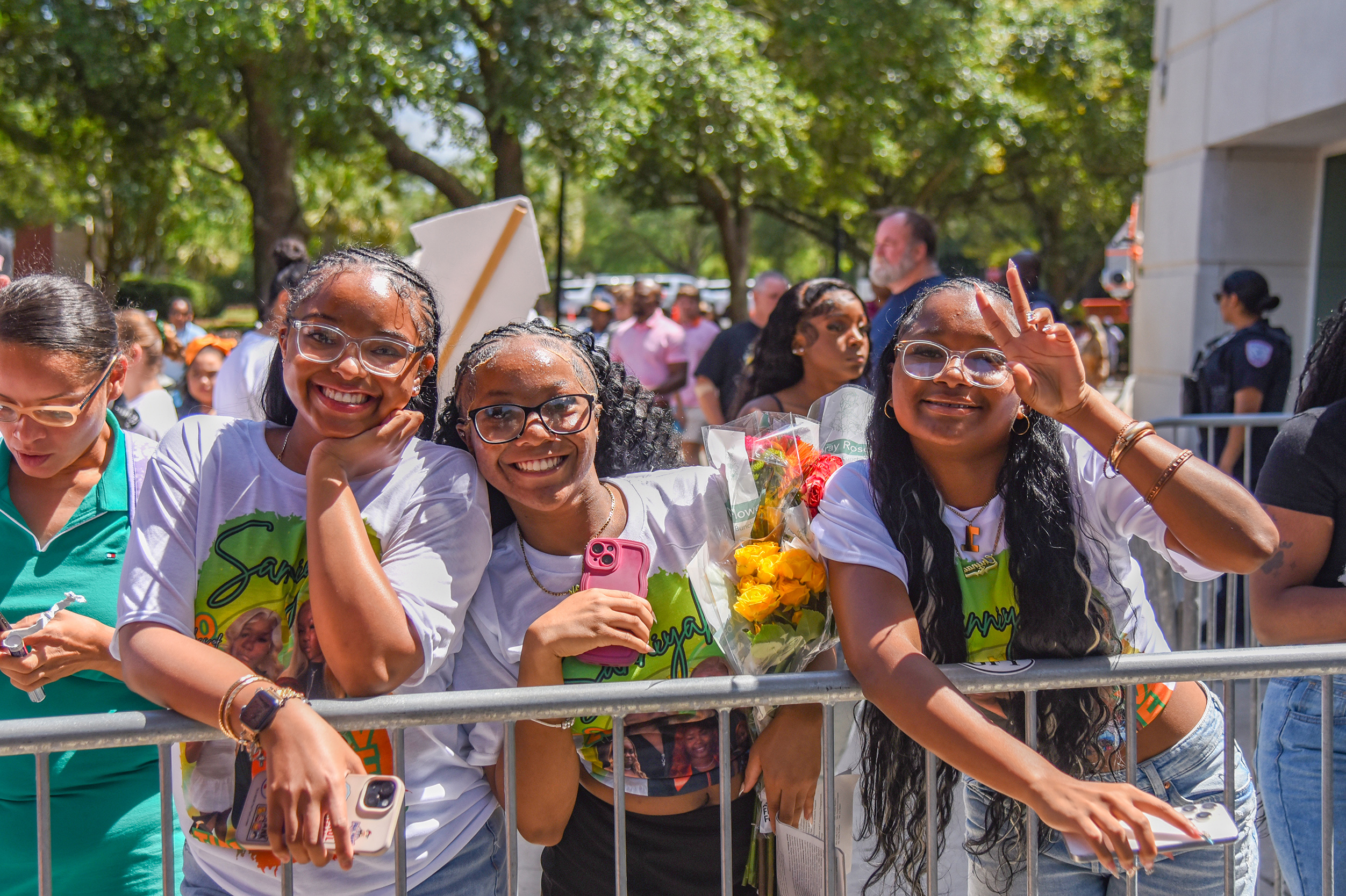 Four women lean on a fence, two giving peace signs. They wear white t-shirts. In the background, people stand behind a fence.