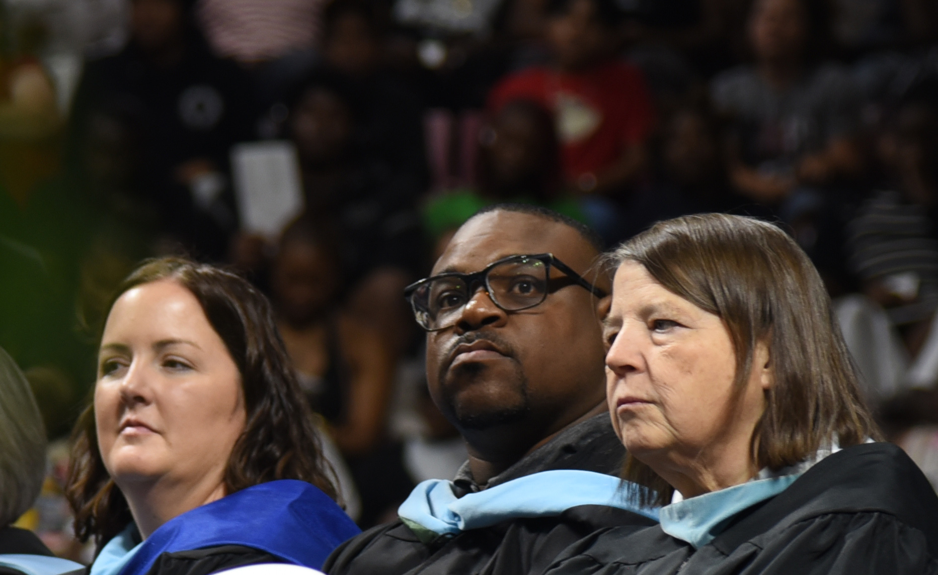Three people in graduation gowns watch the stage at a ceremony, with audience members in the background.