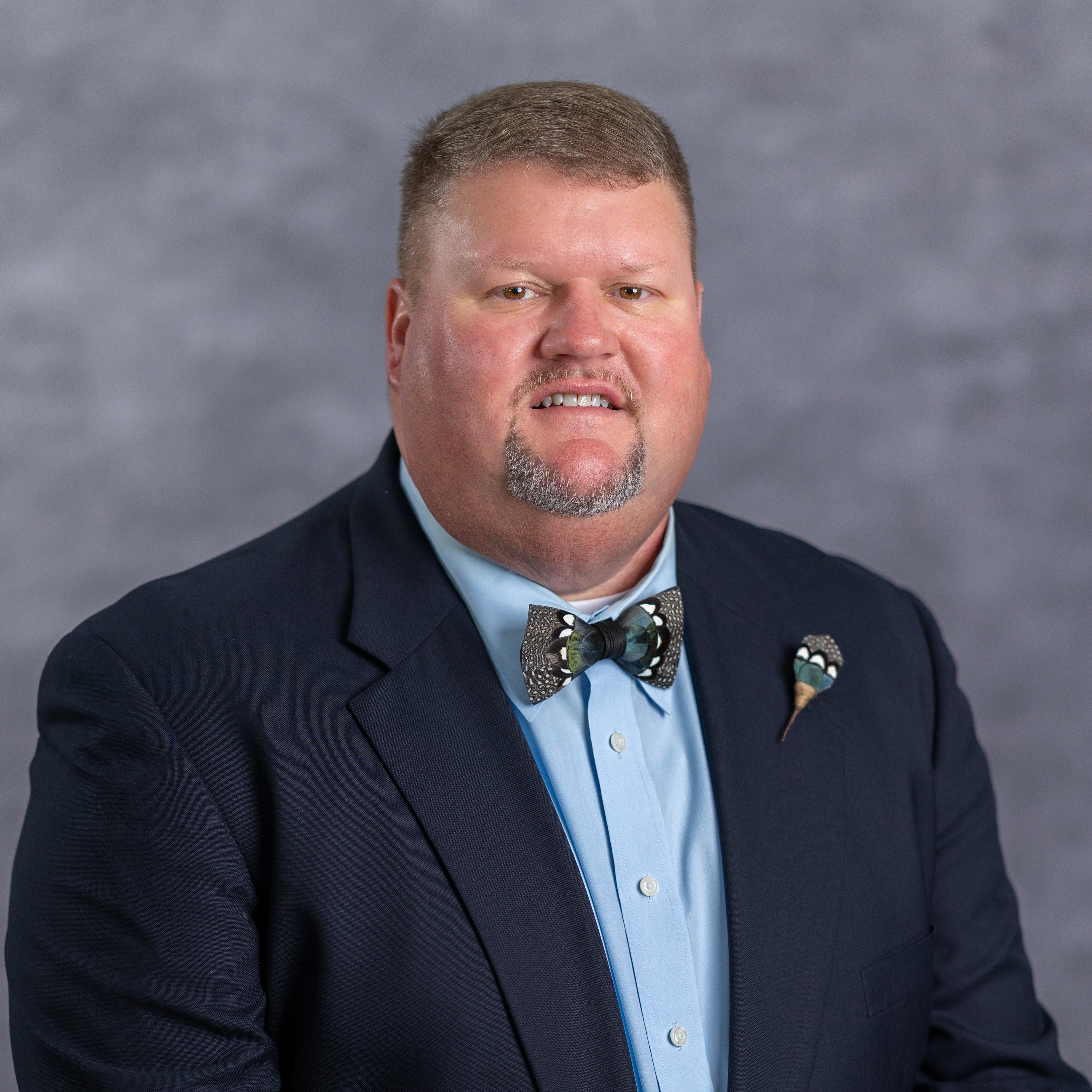 A man with a beard in a blue shirt and dark suit jacket, wearing a bow tie with a pin.