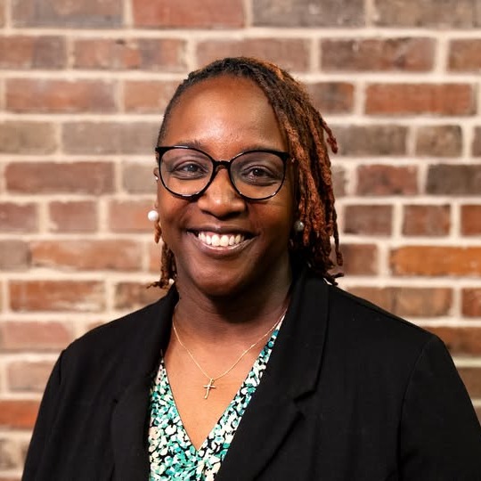 A woman with short hair and glasses stands smiling against a brick wall. She wears a black jacket and a green and blue top.