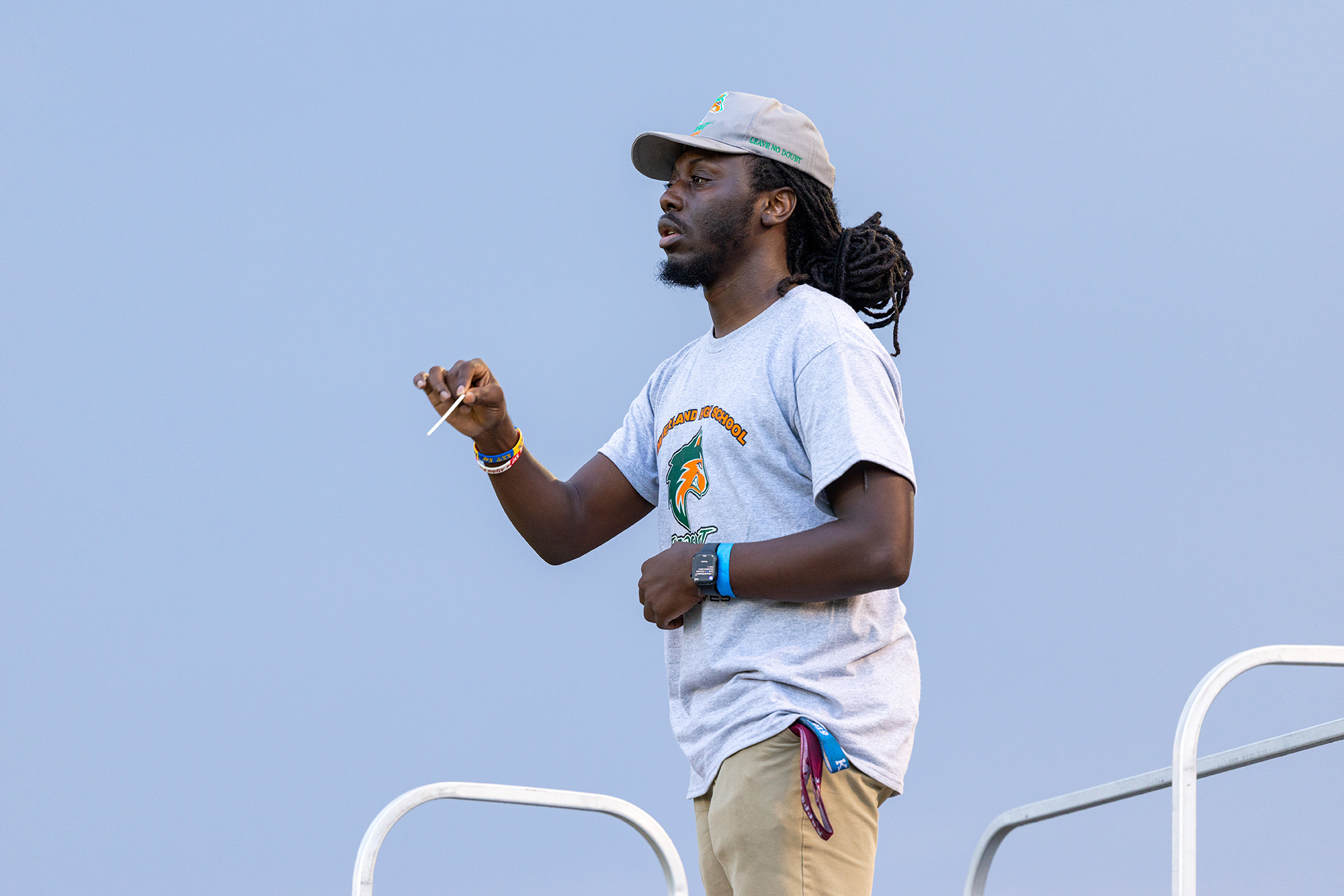 Person with dreadlocks, standing on a metal platform, wearing a white shirt and beige pants, and holding a whistle.