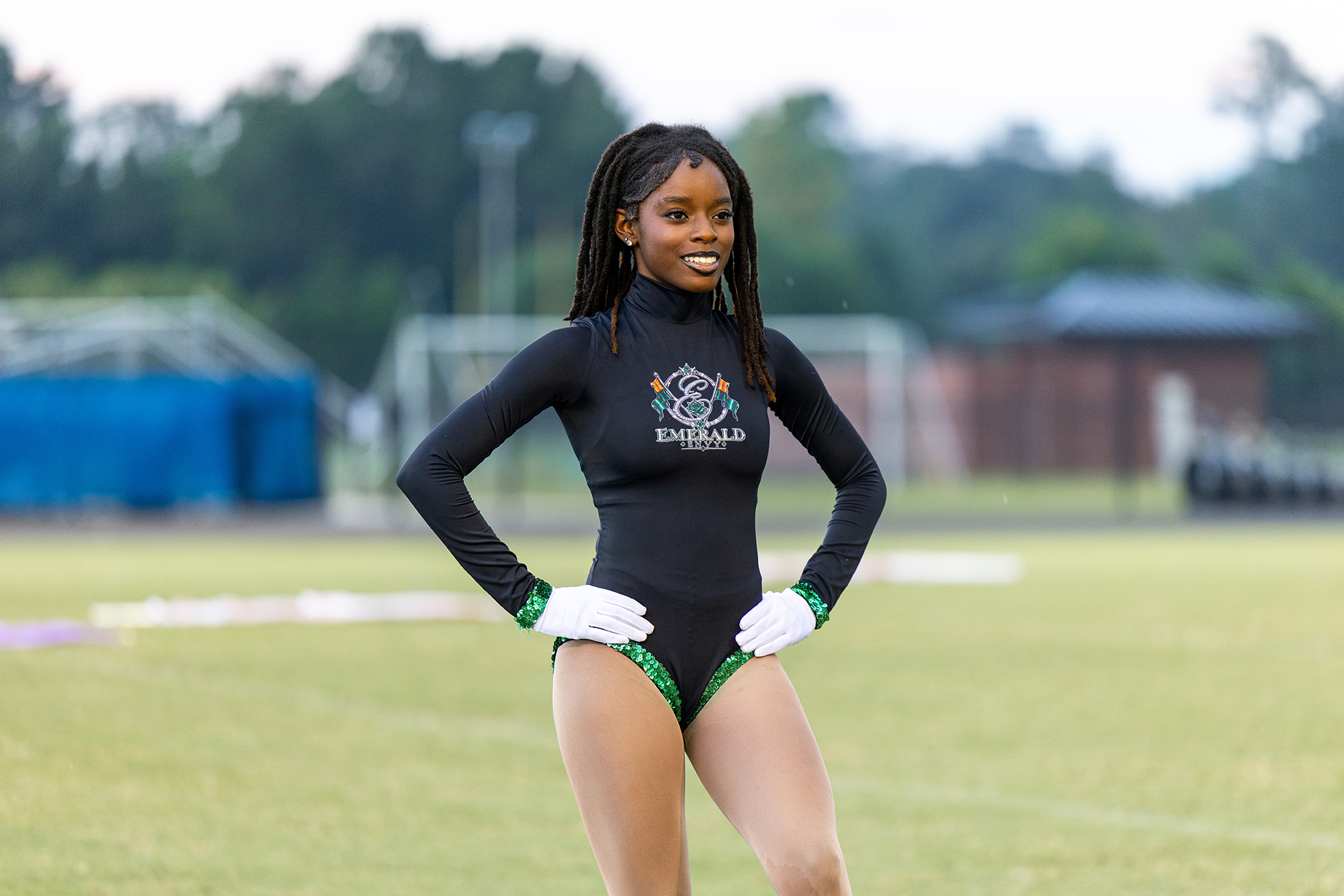Person in black leotard with green trim, white gloves, and high ponytail stands on a grass field.