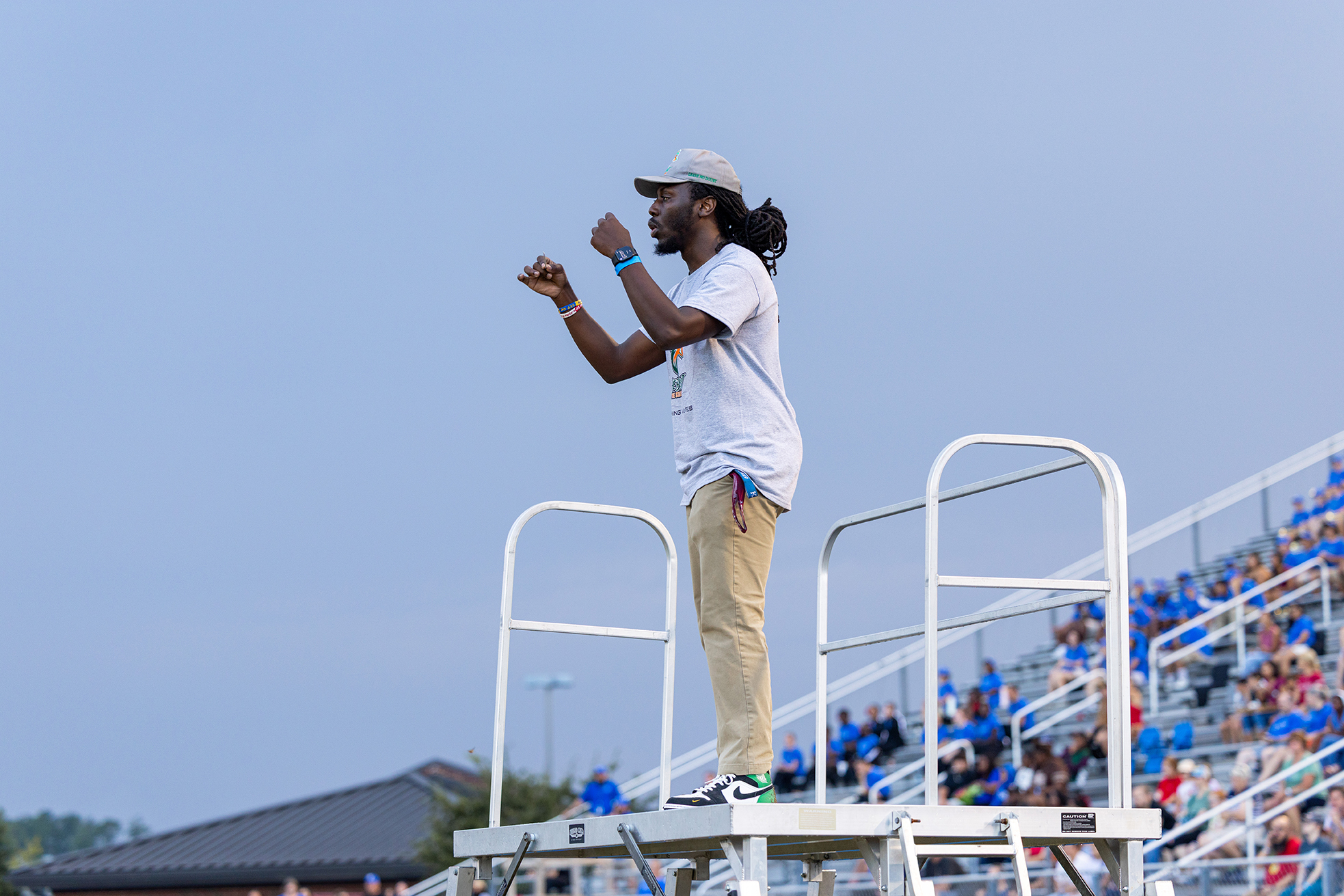 A person stands on a raised platform, wearing a white t-shirt and khaki pants. He gestures with one hand, surrounded by a crowd.