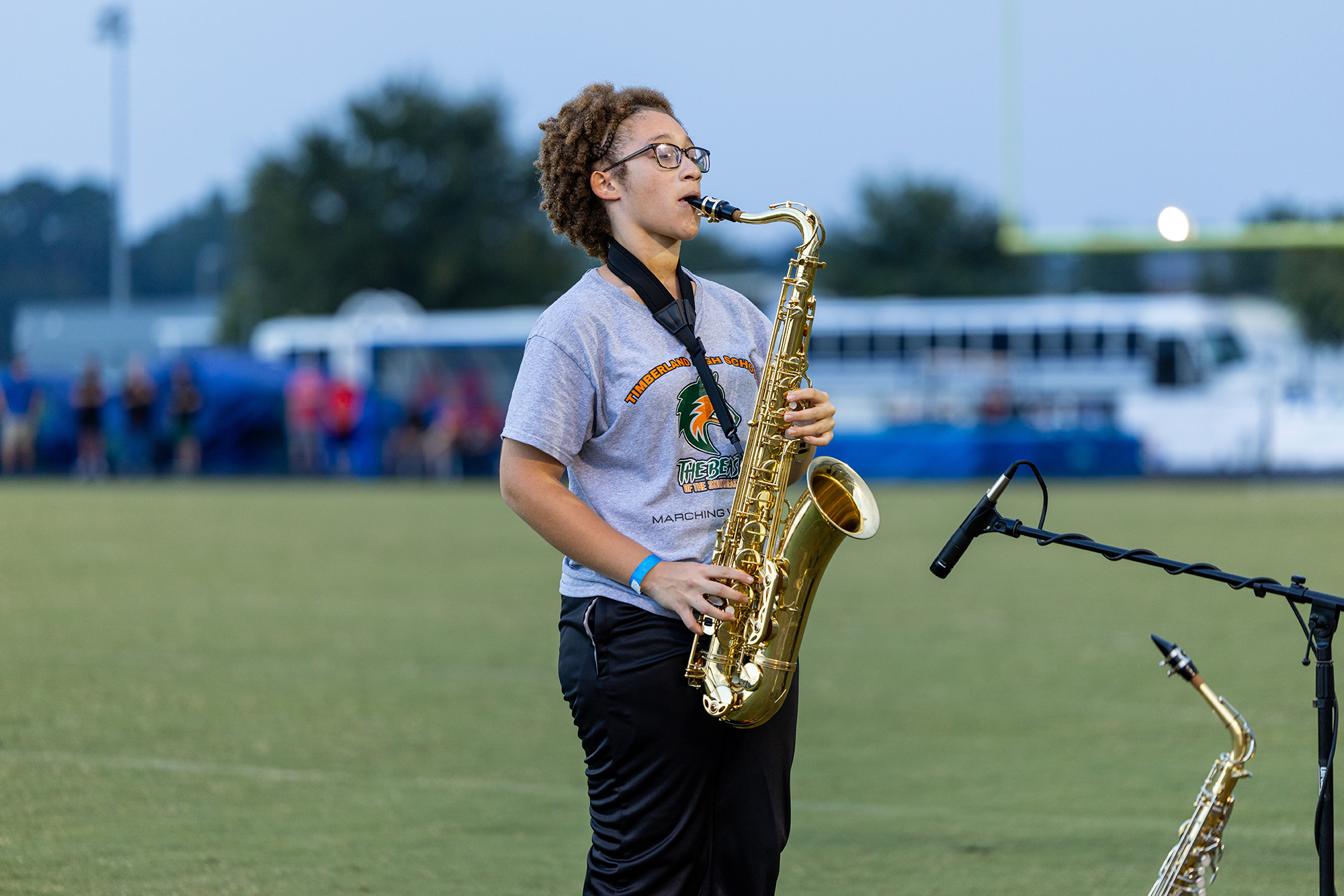 Person playing saxophone outdoors on a football field. Another saxophone stands nearby. A microphone on a stand and people in the background.