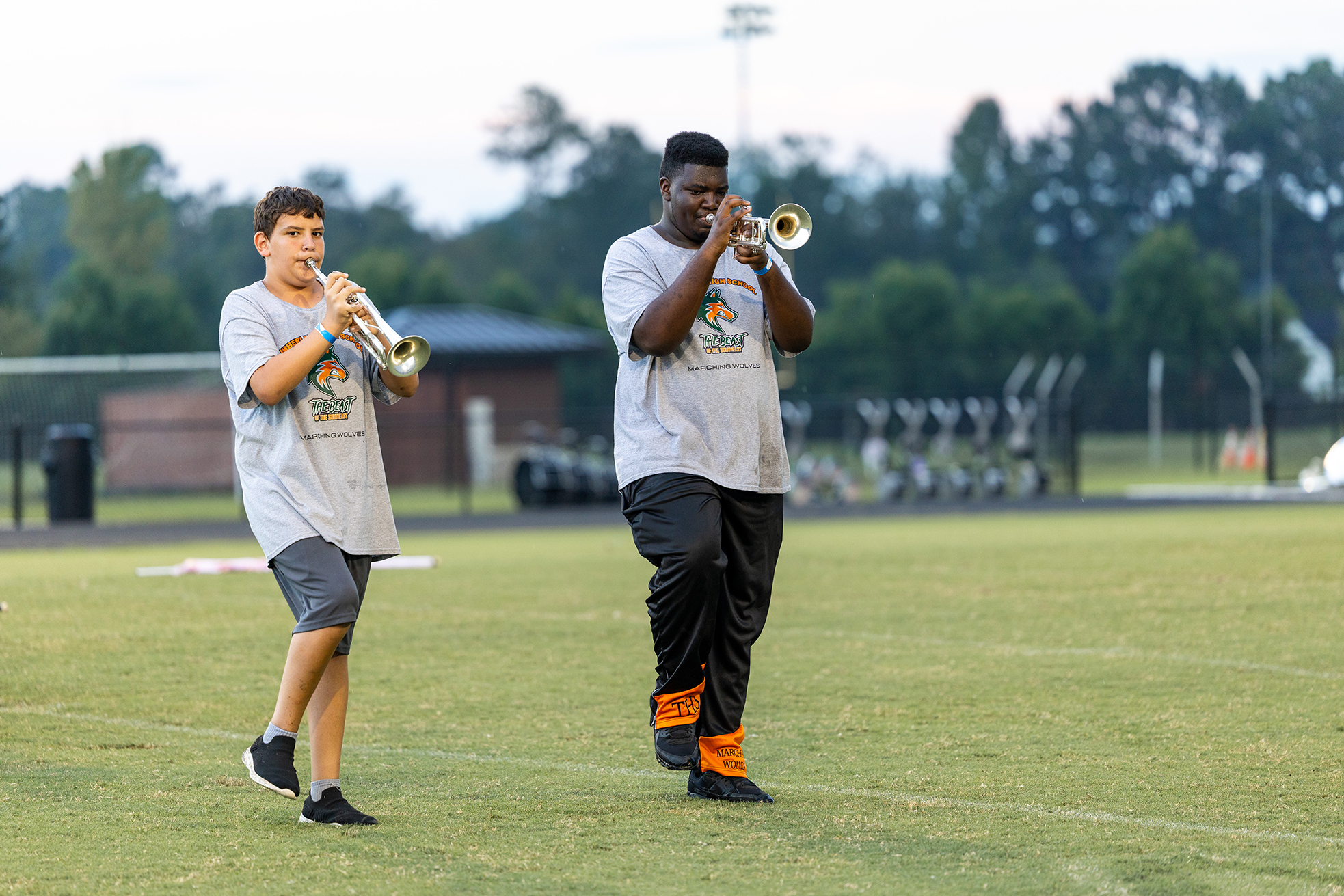 Two people in gray t-shirts and shorts play trumpets on a grass field with trees in the background.