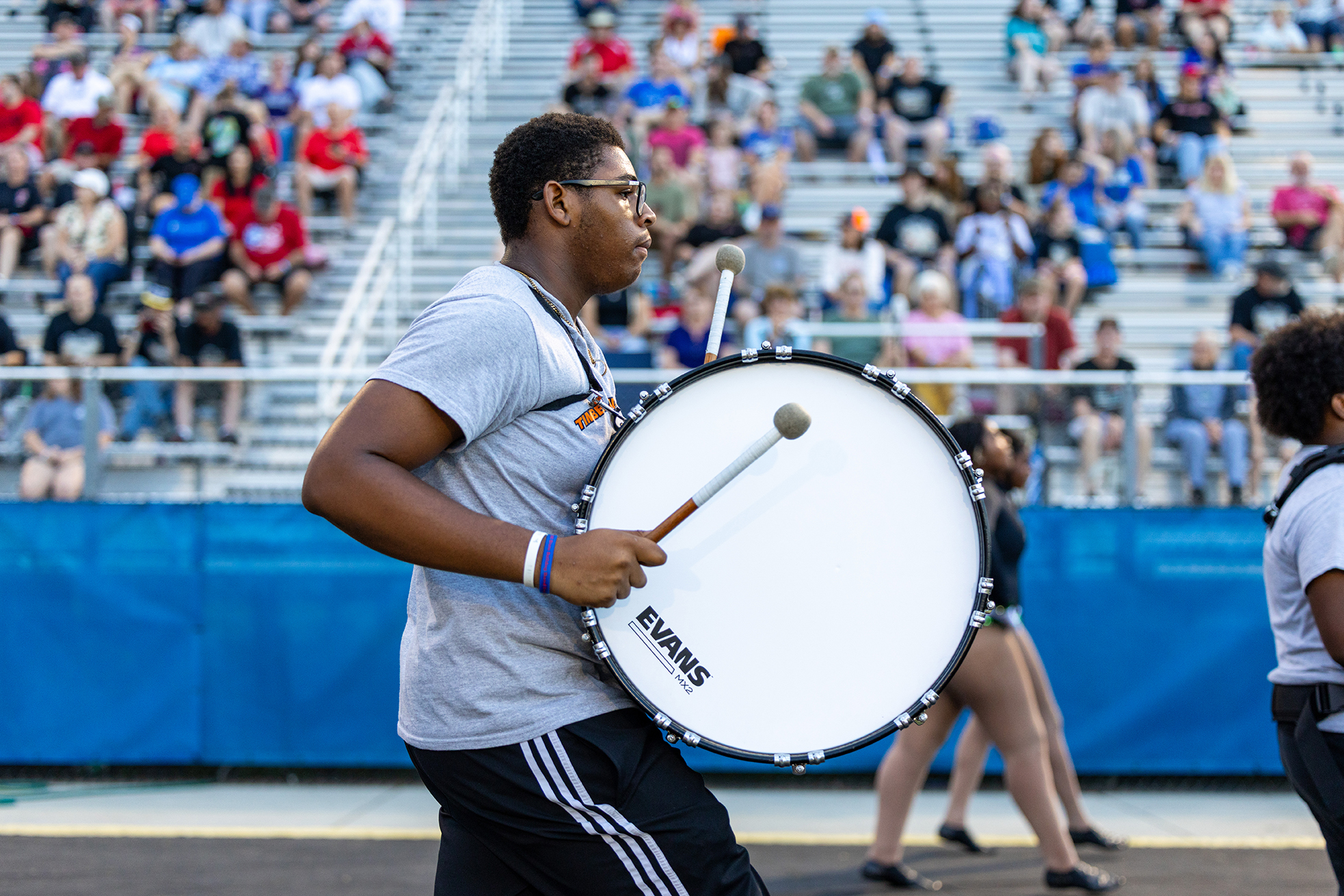 A drummer with a large drum performs in front of a stadium with an audience.